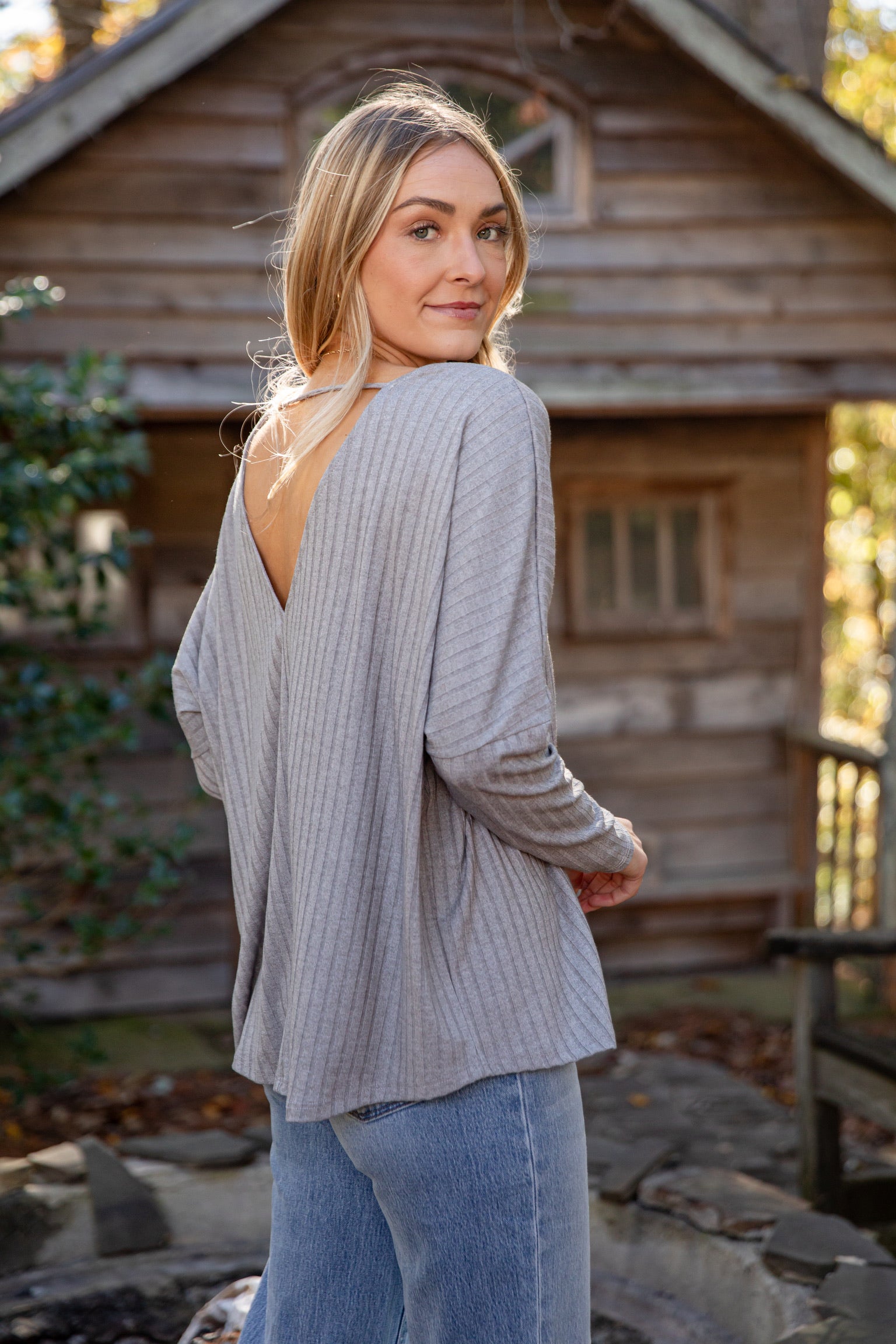 Woman wearing a light gray open-back cardigan and blue jeans standing in front of a wooden cabin.