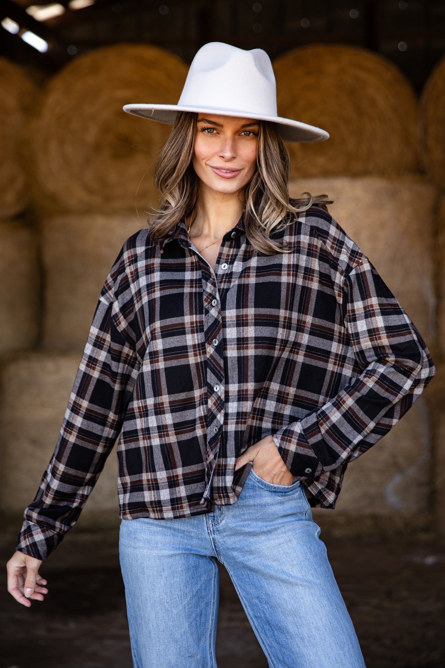 Woman wearing a plaid shirt, jeans, and a white hat in front of hay bales.