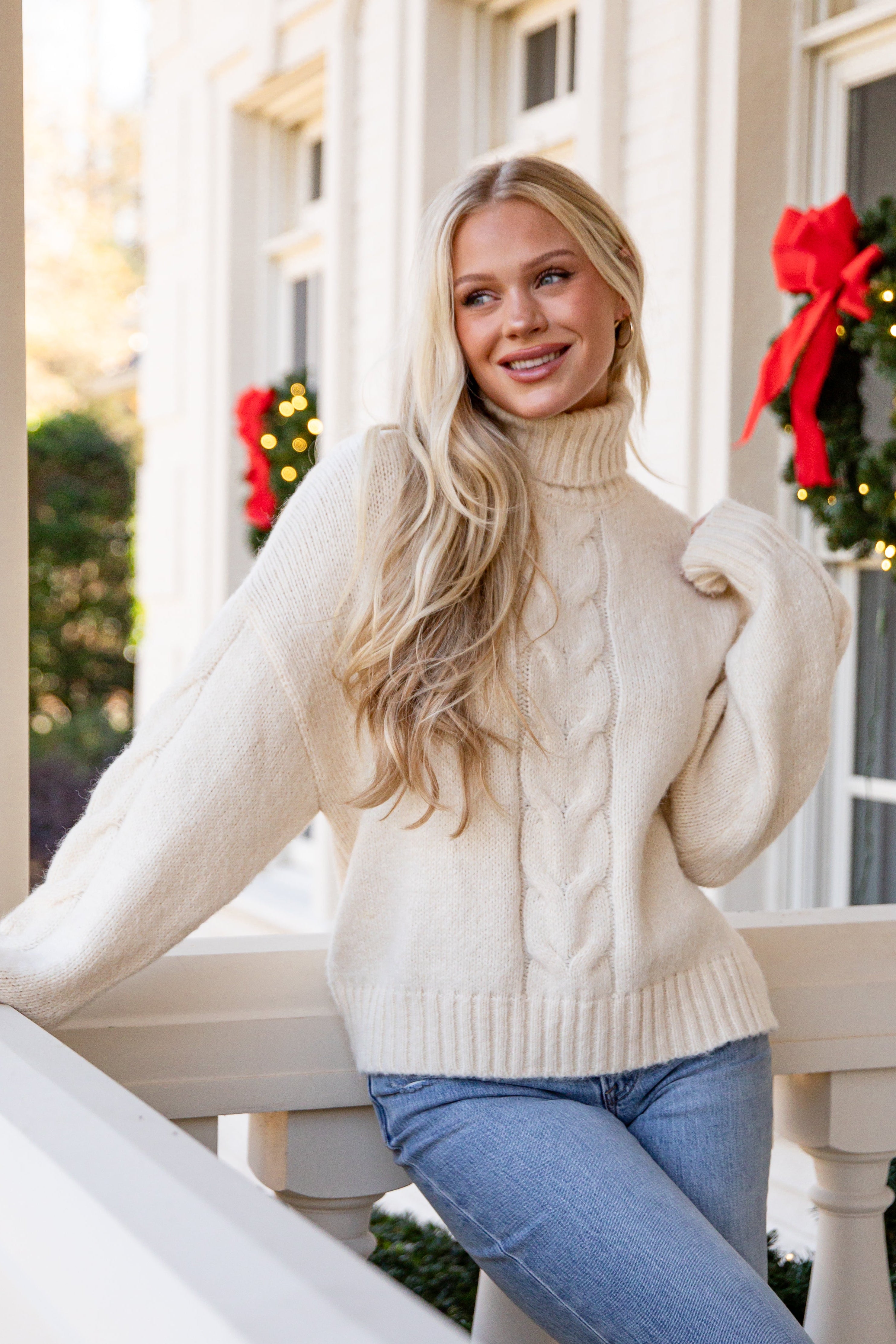 Woman in a cream sweater and blue jeans standing on a balcony with Christmas decorations.