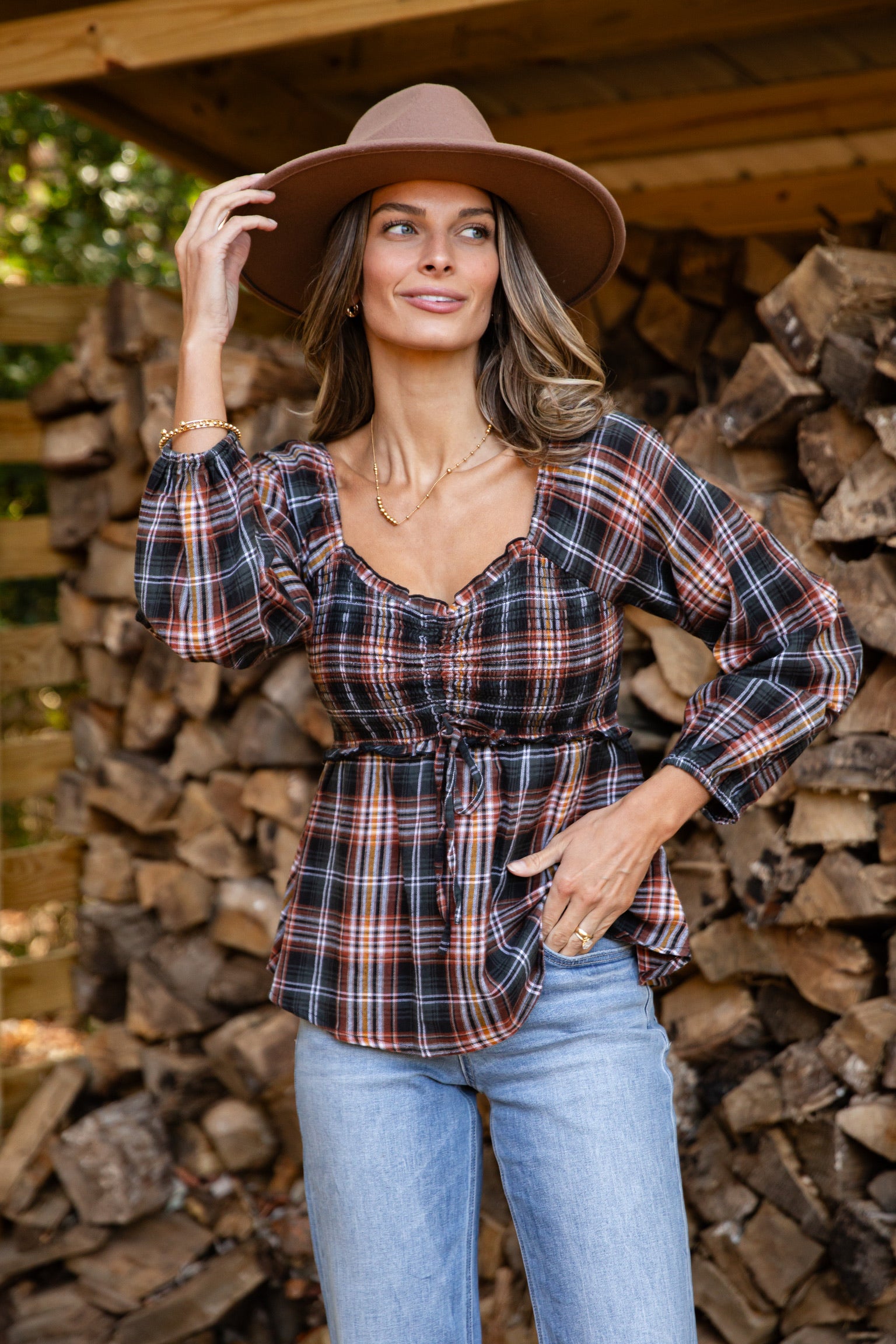 Woman wearing a plaid shirt and wide-brimmed hat standing in front of stacked firewood.