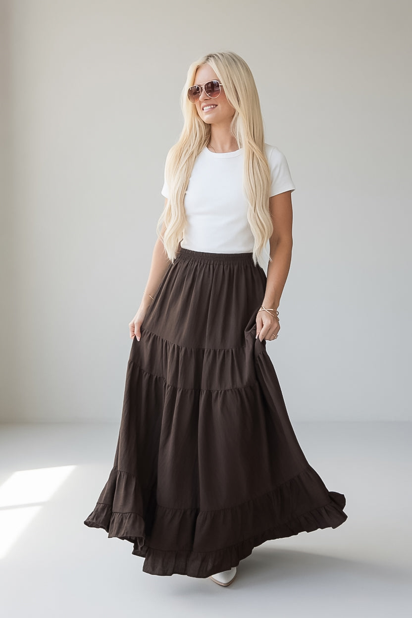 A blonde woman wearing a white short-sleeve t-shirt and a long, dark brown tiered maxi skirt with pockets, standing in a bright, minimalist studio.