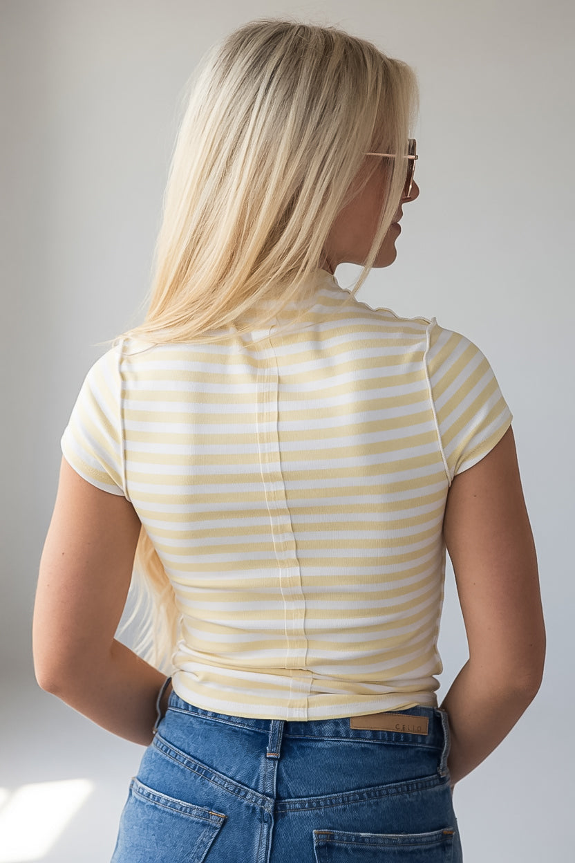 A woman with long blonde hair is shown from behind wearing a pale yellow and white horizontally striped short-sleeve top and blue jeans against a light gray background.