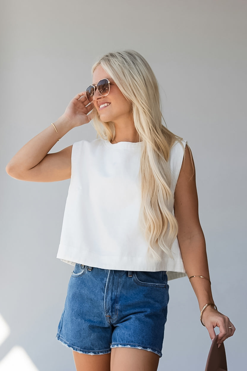 A blonde woman wearing a sleeveless white crop top and blue denim shorts, posing against a light gray background.