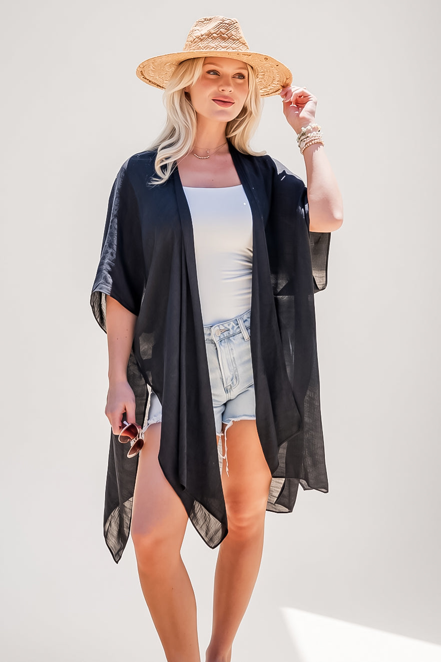 A woman modeling a lightweight black open-front kimono cover-up over a white tank top and denim shorts, wearing a straw hat against a plain light background.