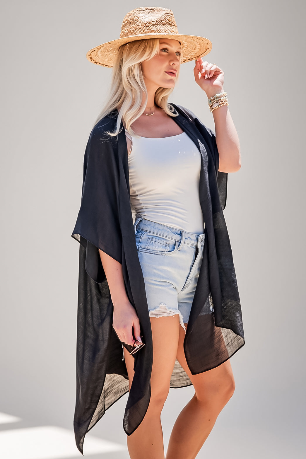 A woman modeling a sheer black open-front kimono-style cover-up over a white tank top and light denim shorts, wearing a straw hat against a plain light gray studio background.