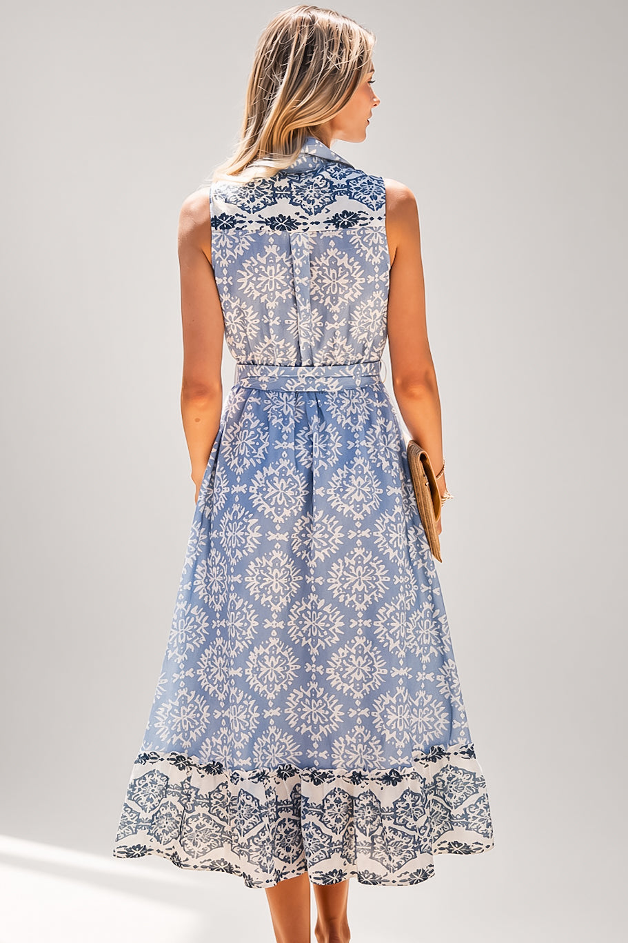 A woman modeling a sleeveless midi dress from the back, featuring a light blue and white damask pattern with black lace trim at the high neckline, waist, and ruffled hem, set against a plain light gray studio background.
