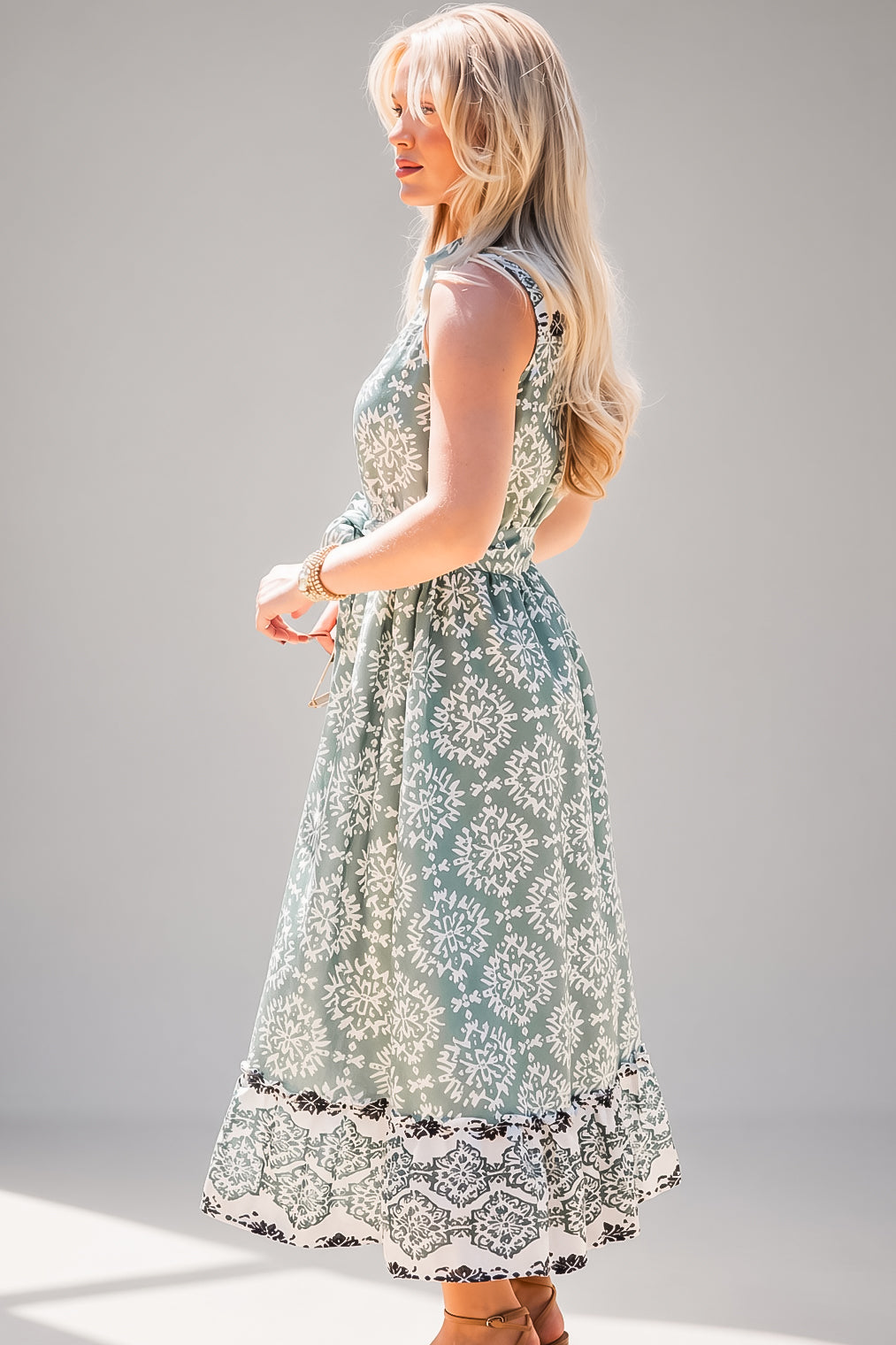 A woman modeling a sleeveless midi dress with a sage green and white floral pattern, standing against a neutral gray studio background.