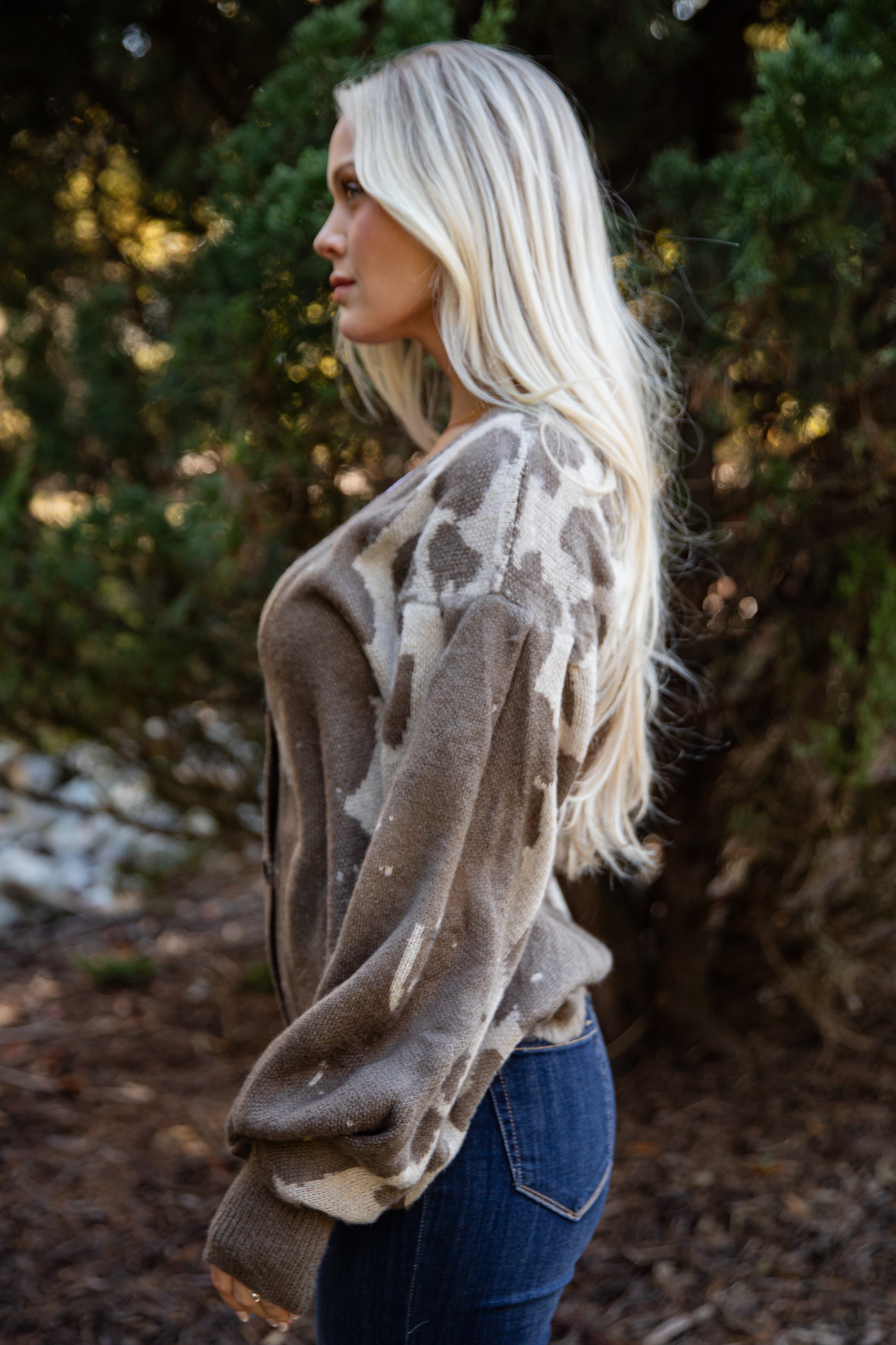 Woman wearing a patterned sweater and jeans standing in a natural setting with trees and ground cover.