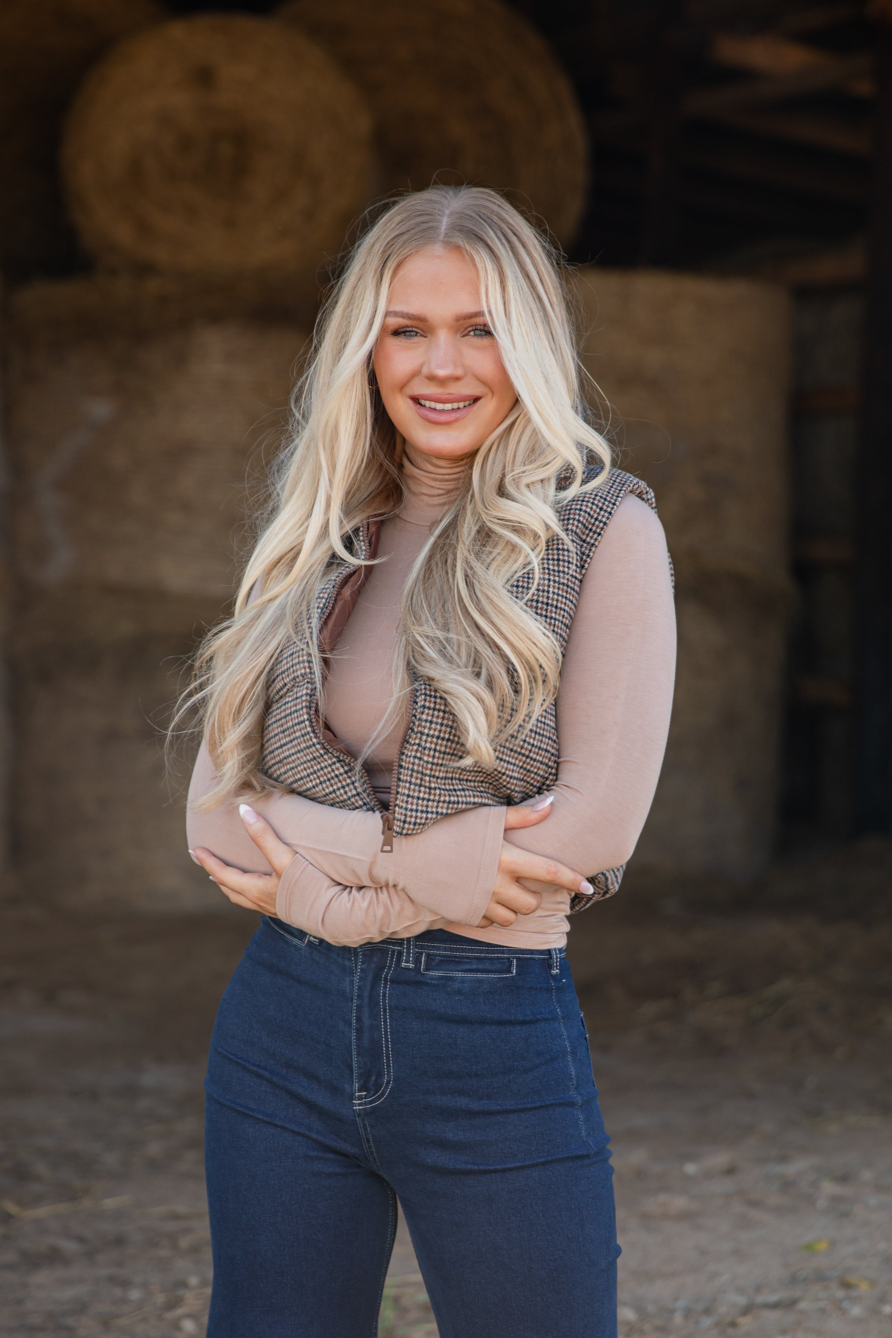 Woman standing in front of hay bales wearing a plaid vest and blue jeans.