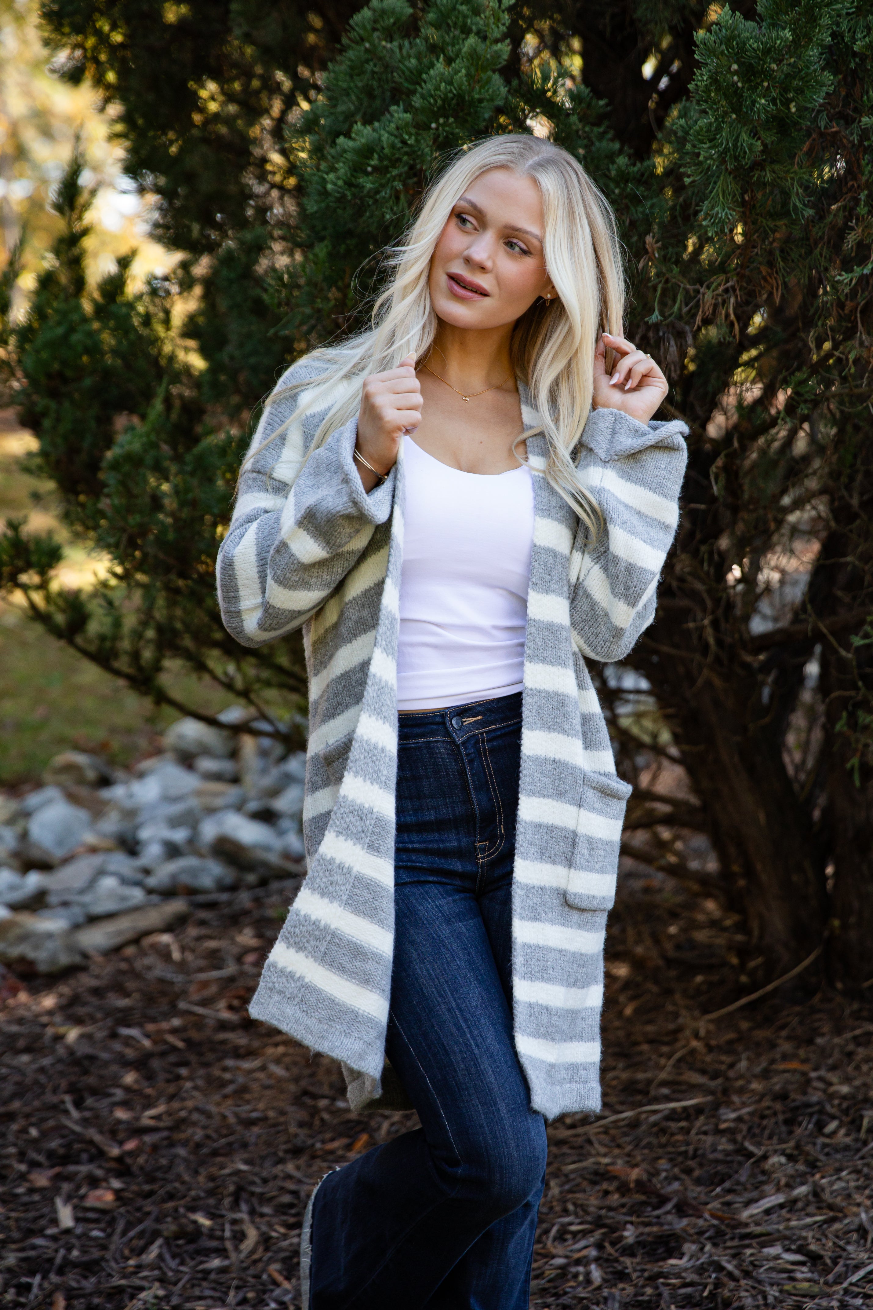 Woman wearing a long, striped cardigan in a natural setting with trees and rocks.