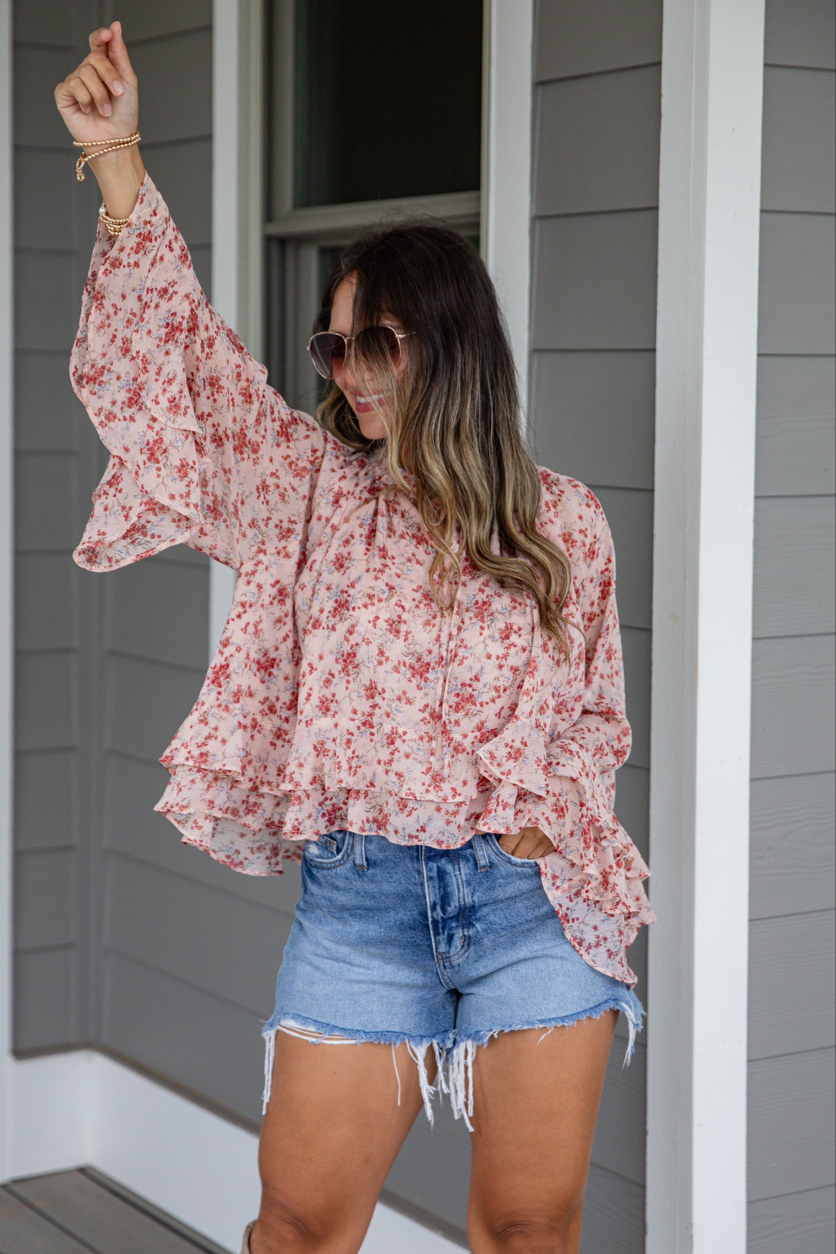 Woman wearing a floral blouse and denim shorts standing on a porch.