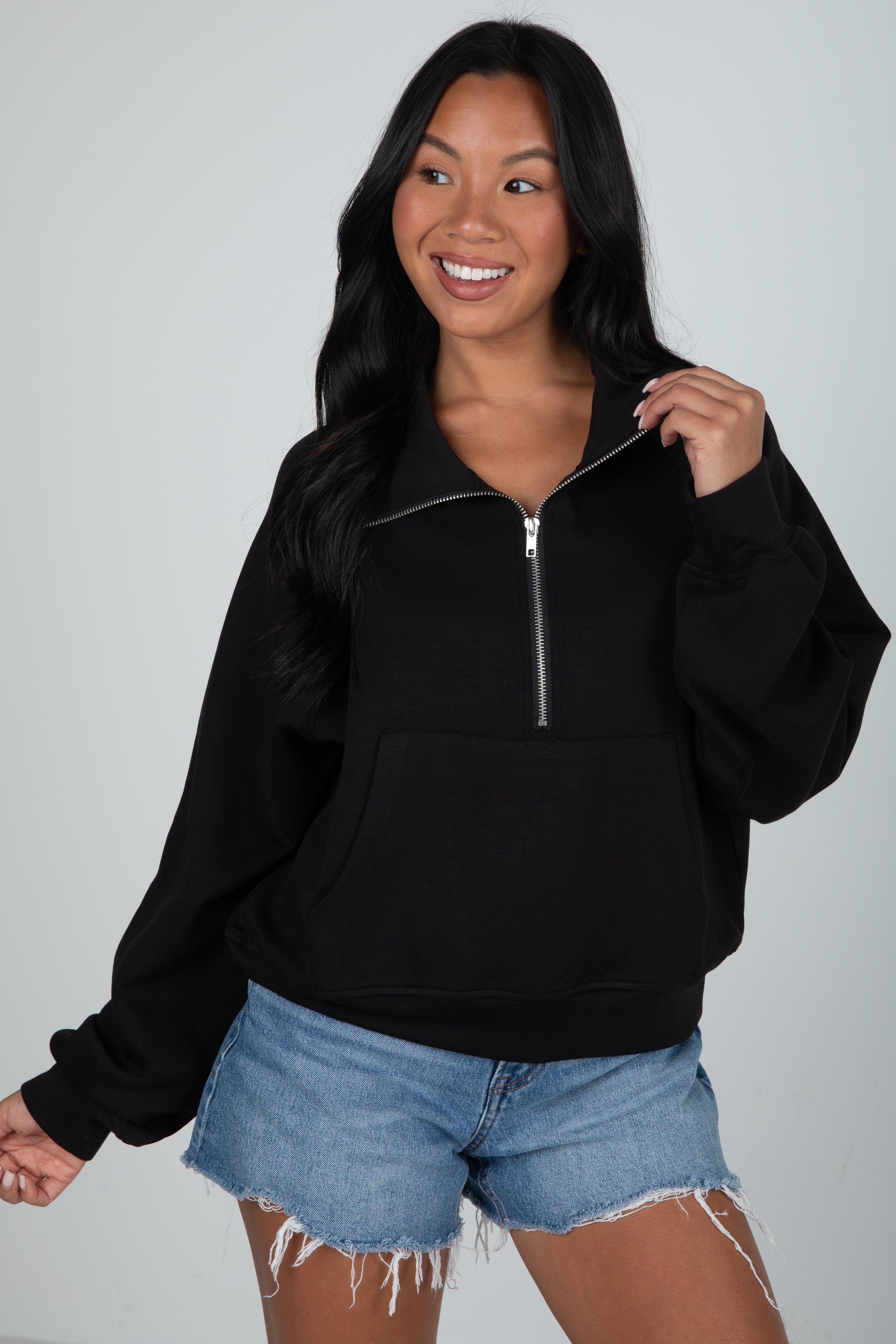 Woman wearing a black half-zip sweatshirt and denim shorts on a gray background