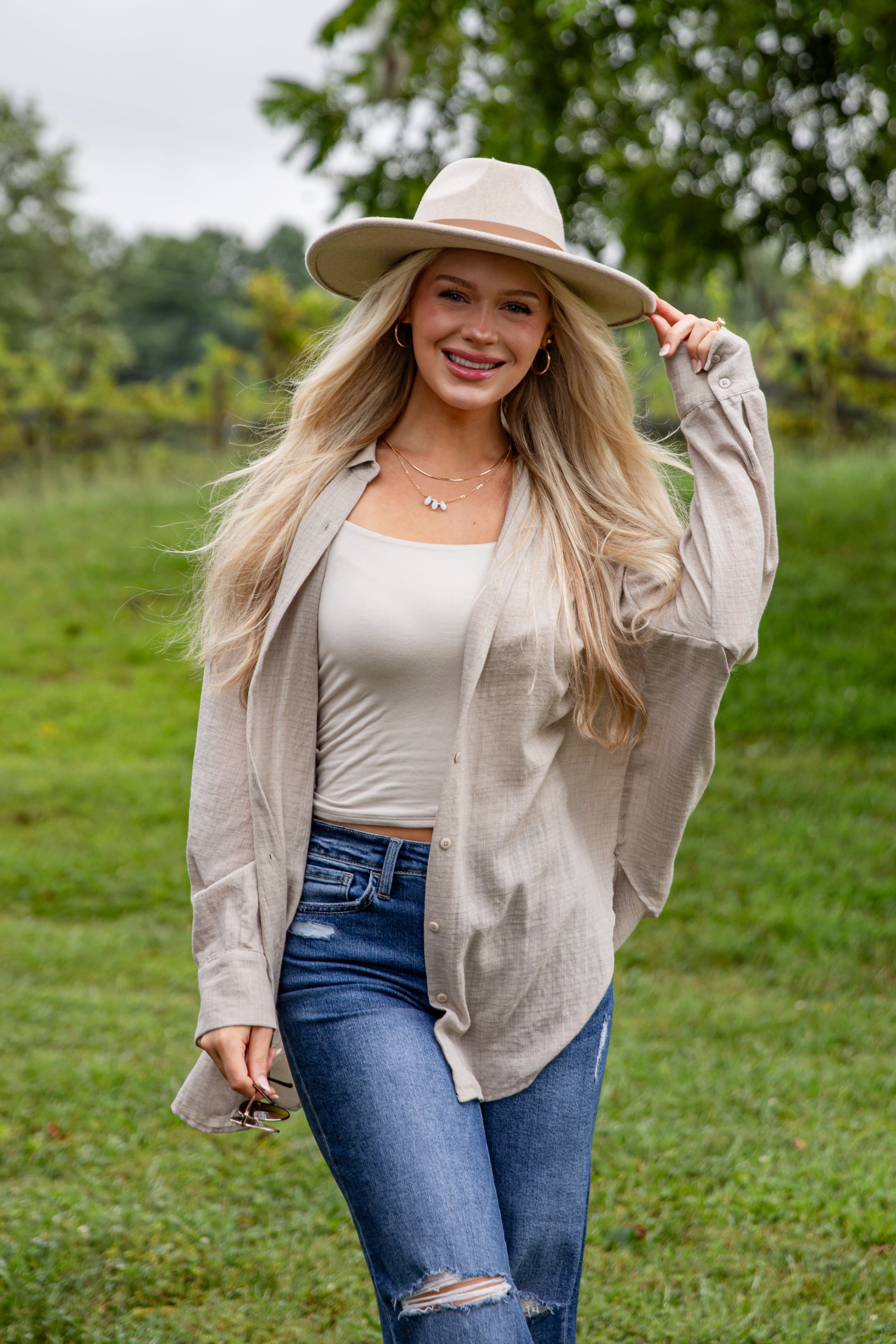Woman wearing a beige hat, beige cardigan, white top, and blue jeans in a grassy outdoor setting.
