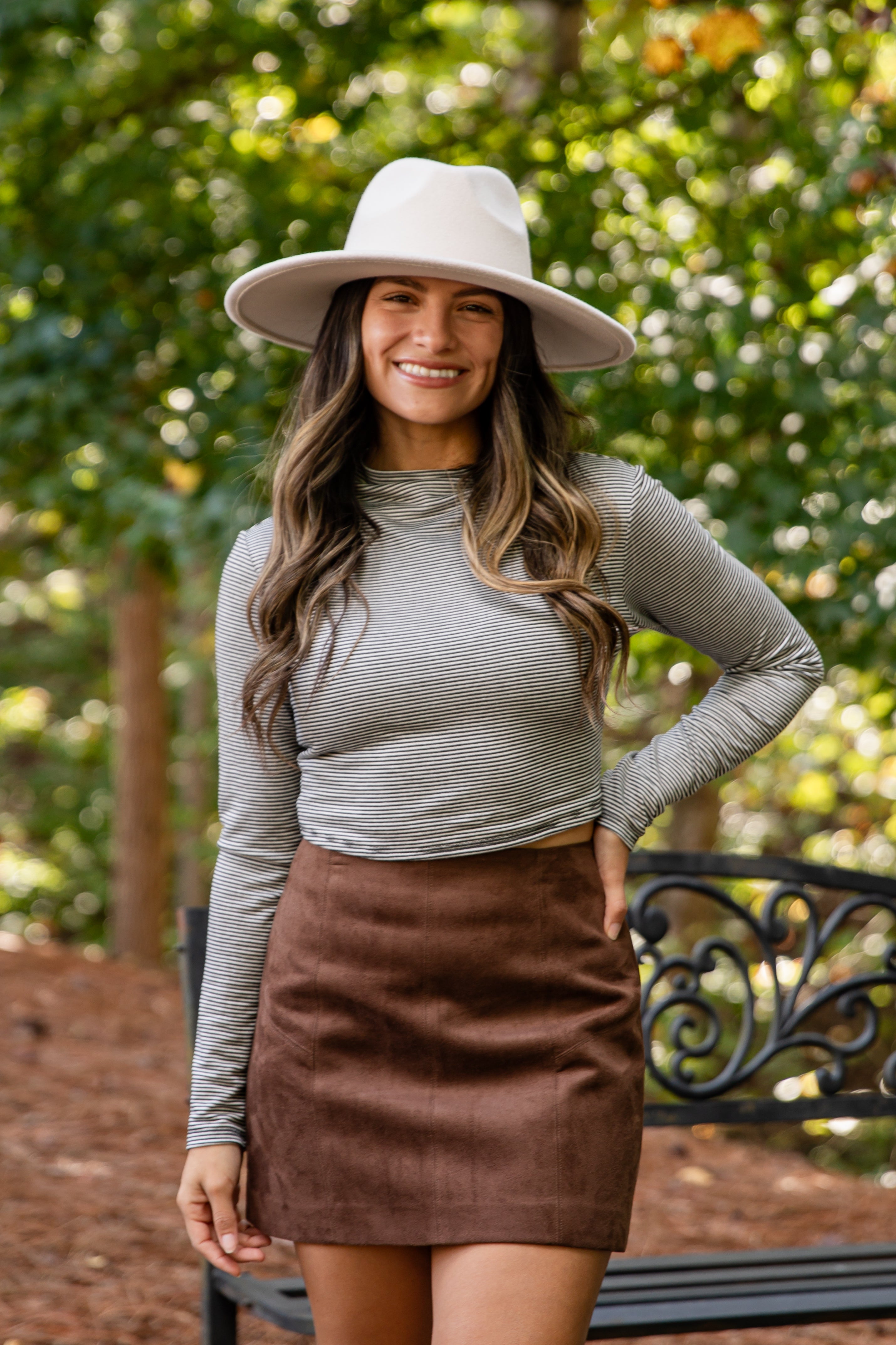 Woman wearing a white hat and brown skirt standing outdoors with greenery in the background