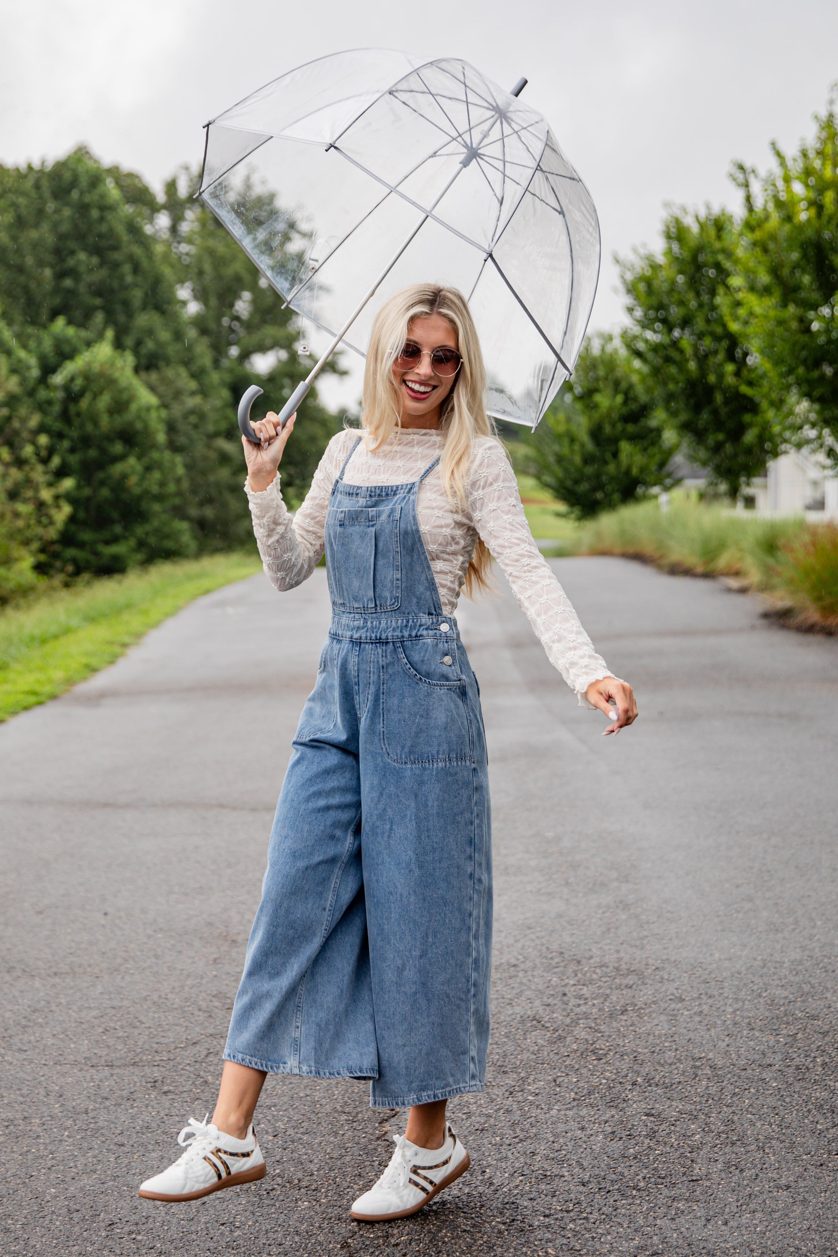 Woman in denim overalls holding a transparent umbrella on a road with trees in the background