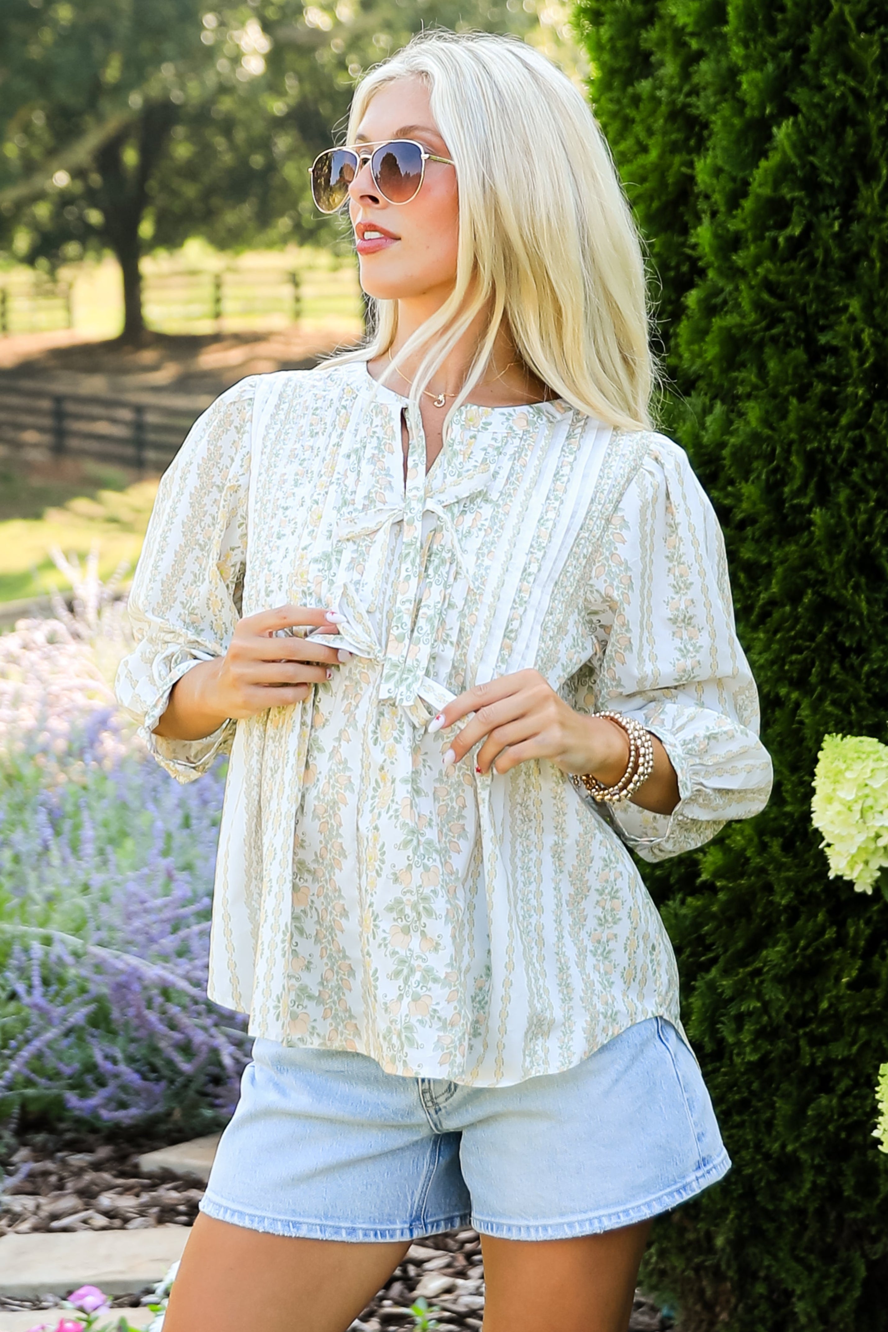 Woman in a white blouse and denim shorts standing outdoors with greenery around