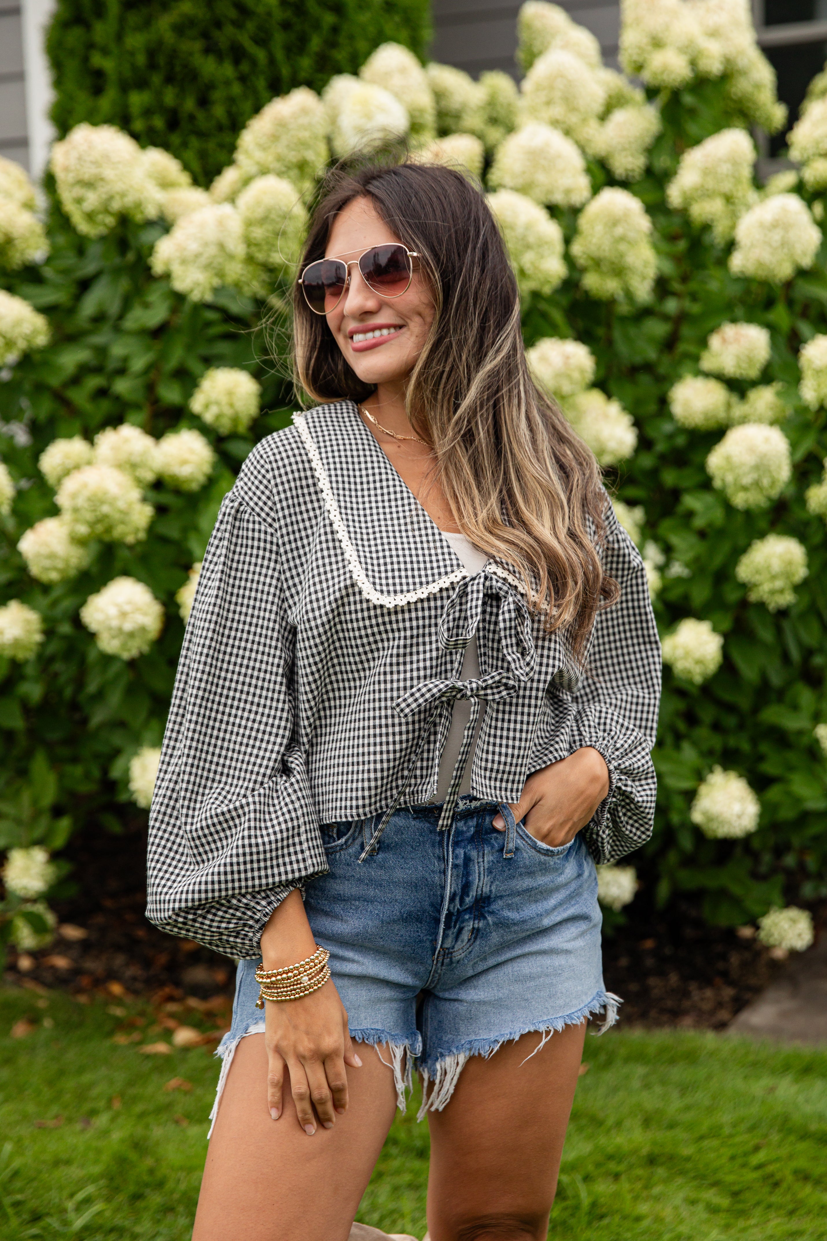 Woman wearing a checkered blouse and denim shorts standing in front of white flowers.