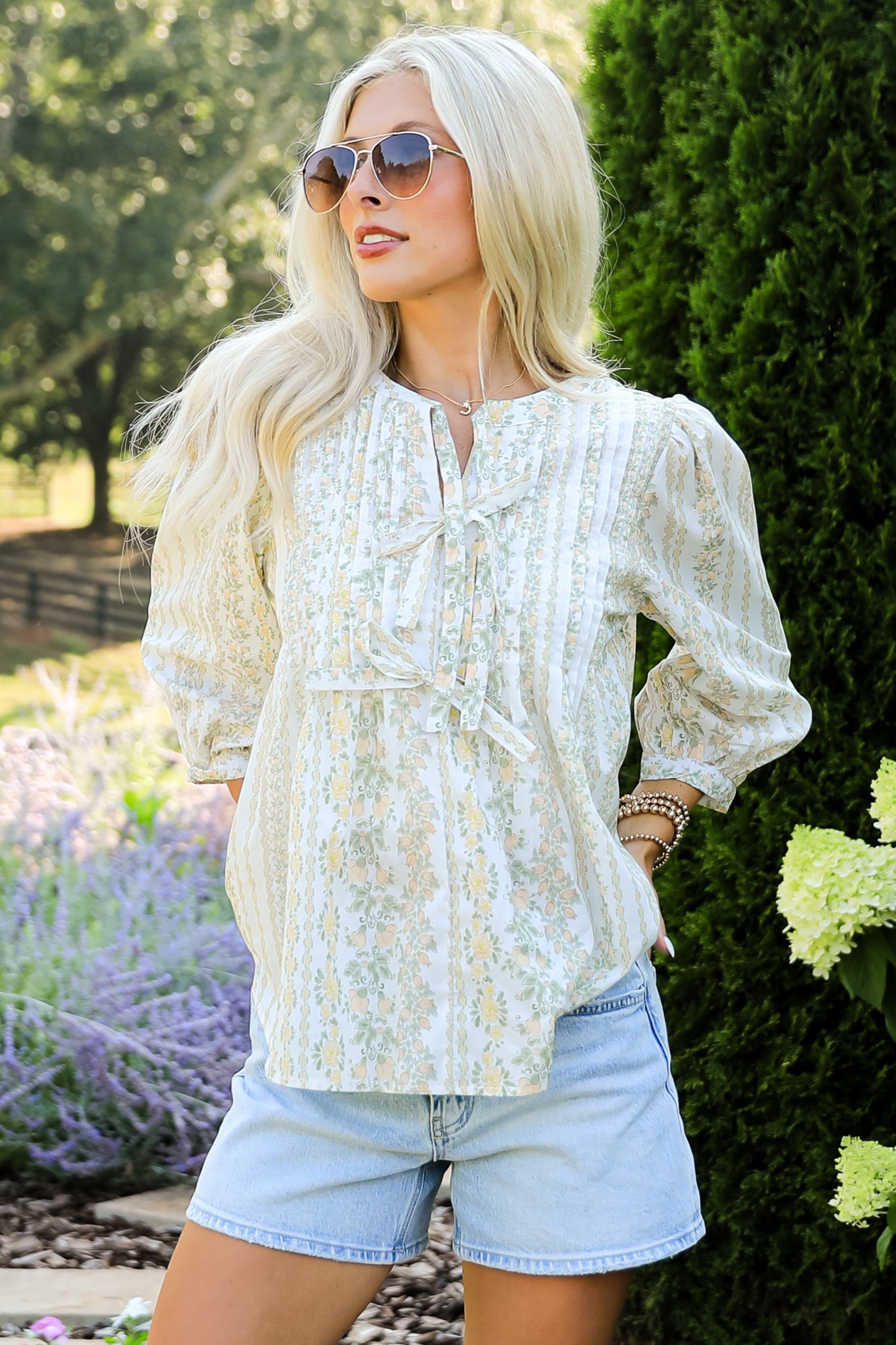 Woman in a garden wearing a white blouse, denim shorts, and brown boots.