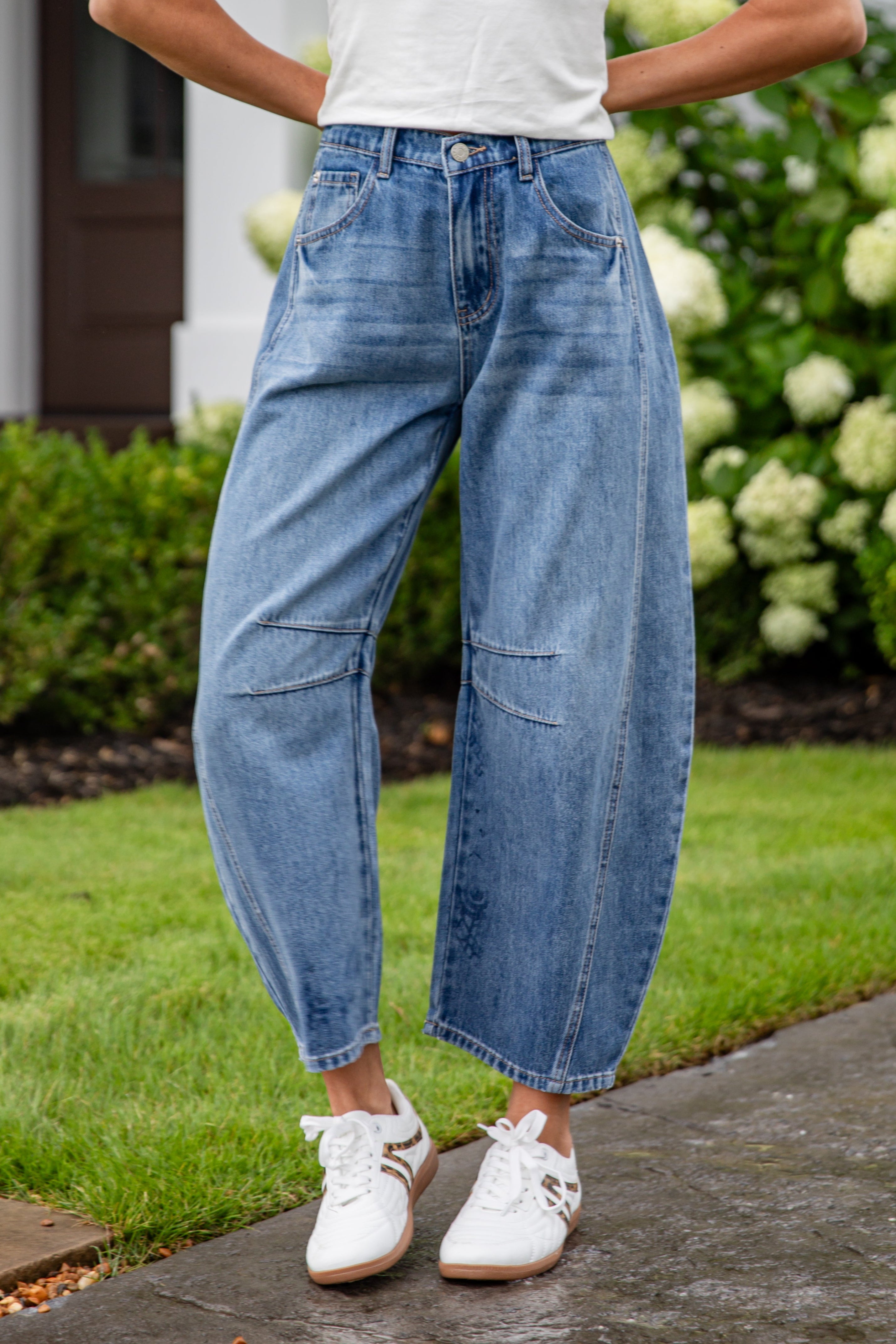 Person wearing a white t-shirt and blue jeans standing on a sidewalk with greenery in the background