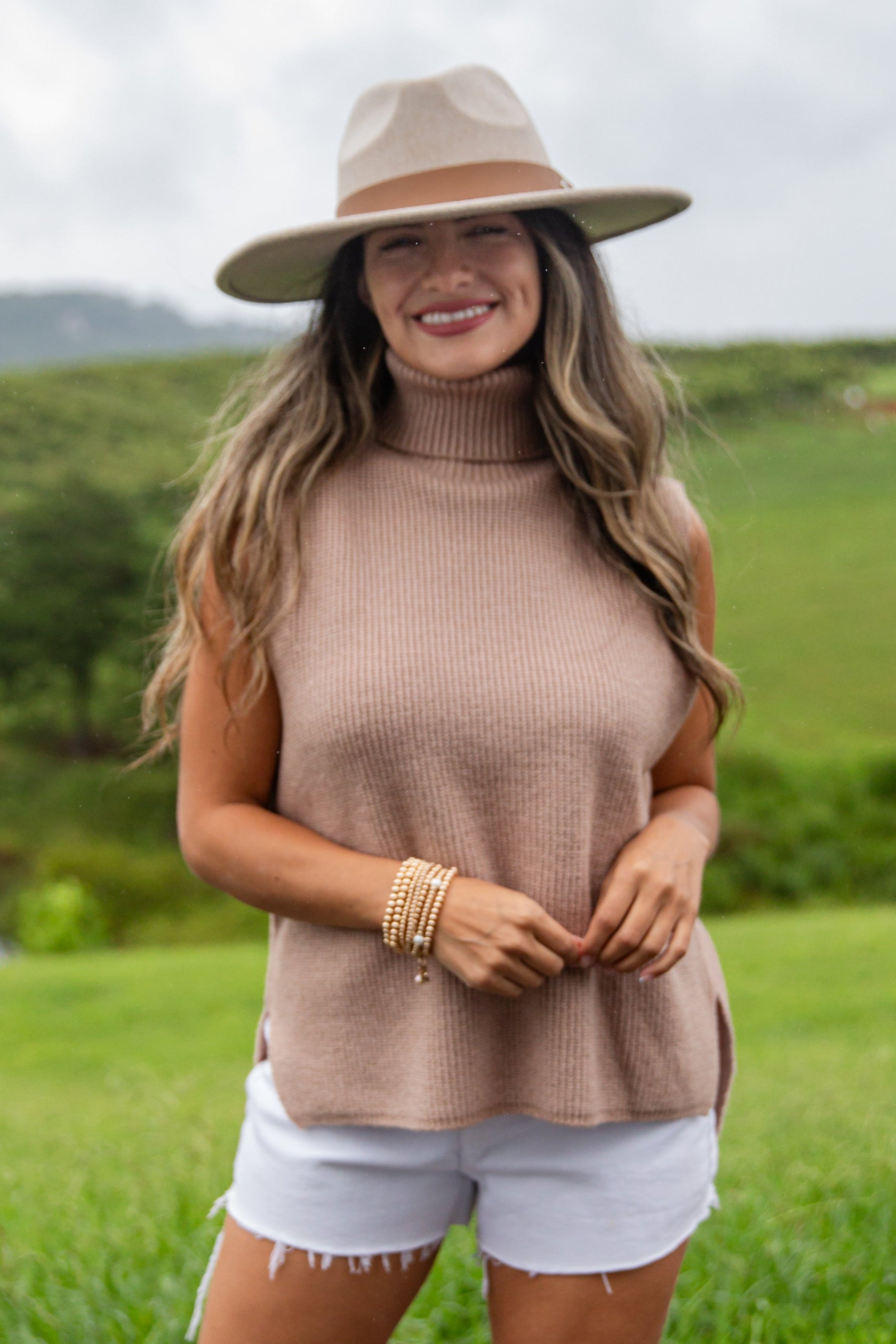 Woman in a sleeveless brown top, white shorts, and beige boots standing outdoors with green fields and a lake in the background.