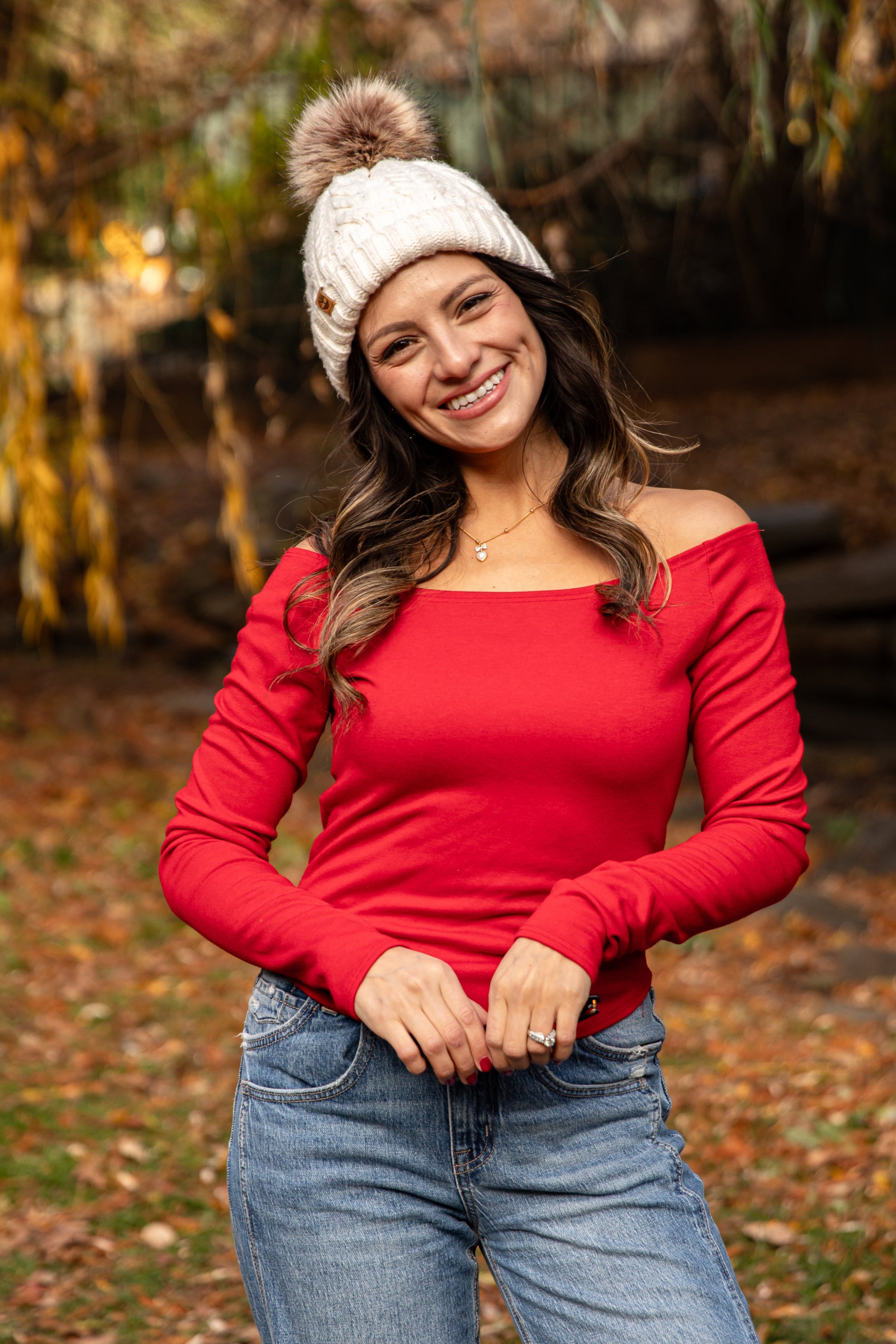 Woman wearing a red top and blue jeans with a white knit hat in an autumn setting.