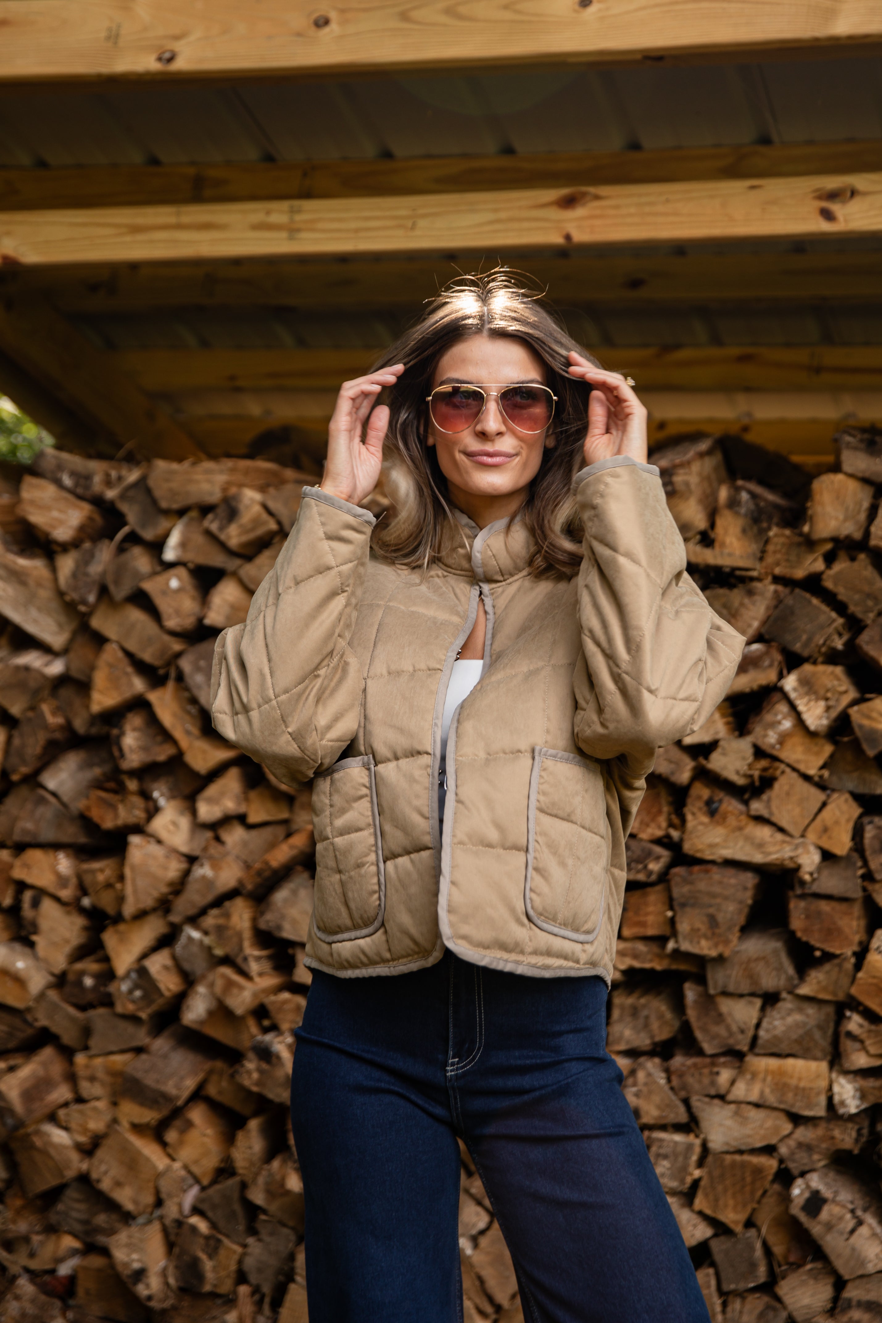 Woman wearing sunglasses and a beige quilted jacket in front of stacked firewood.