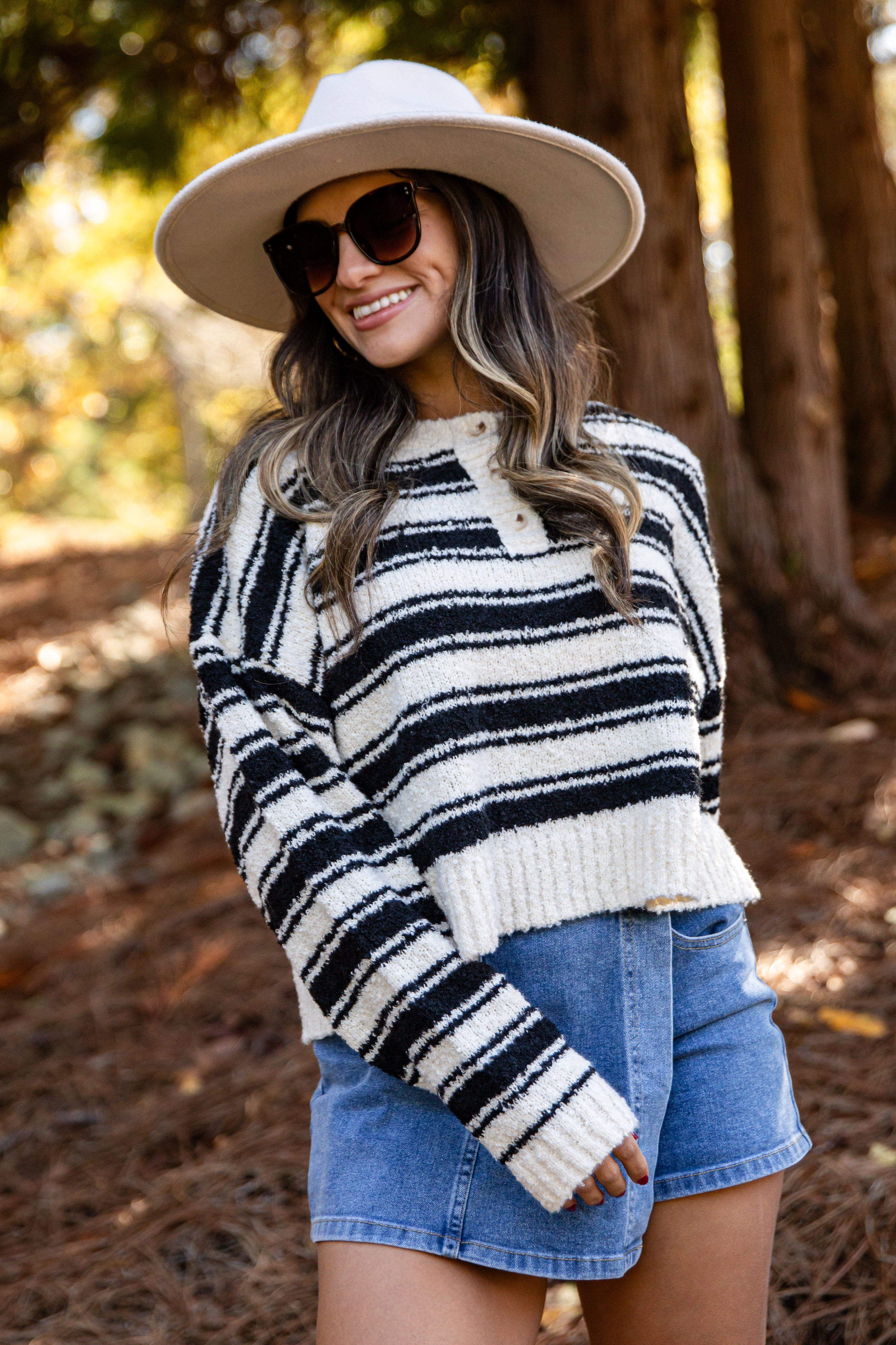 Woman wearing a black and white striped sweater, denim shorts, and a wide-brimmed hat in a forest setting.