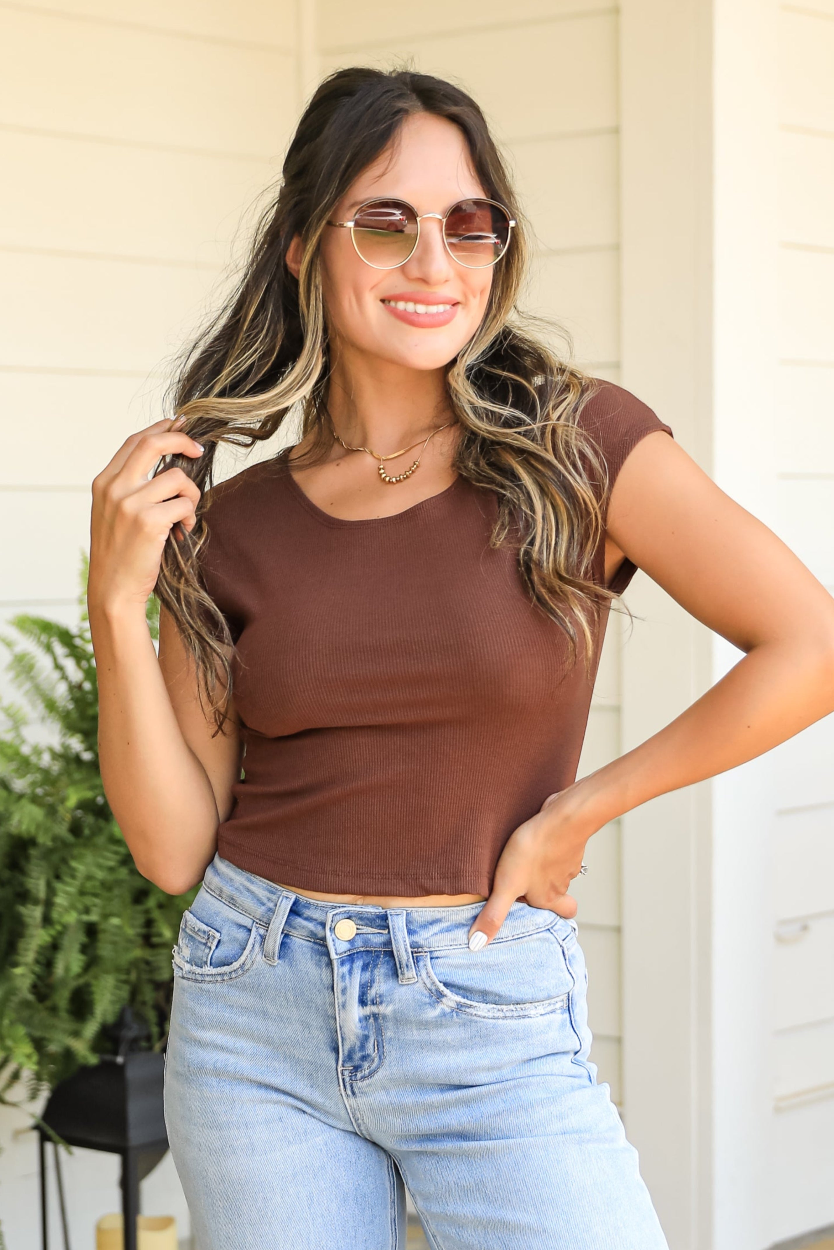 Woman in brown top and light blue jeans standing on a stone patio with plants and a house in the background.
