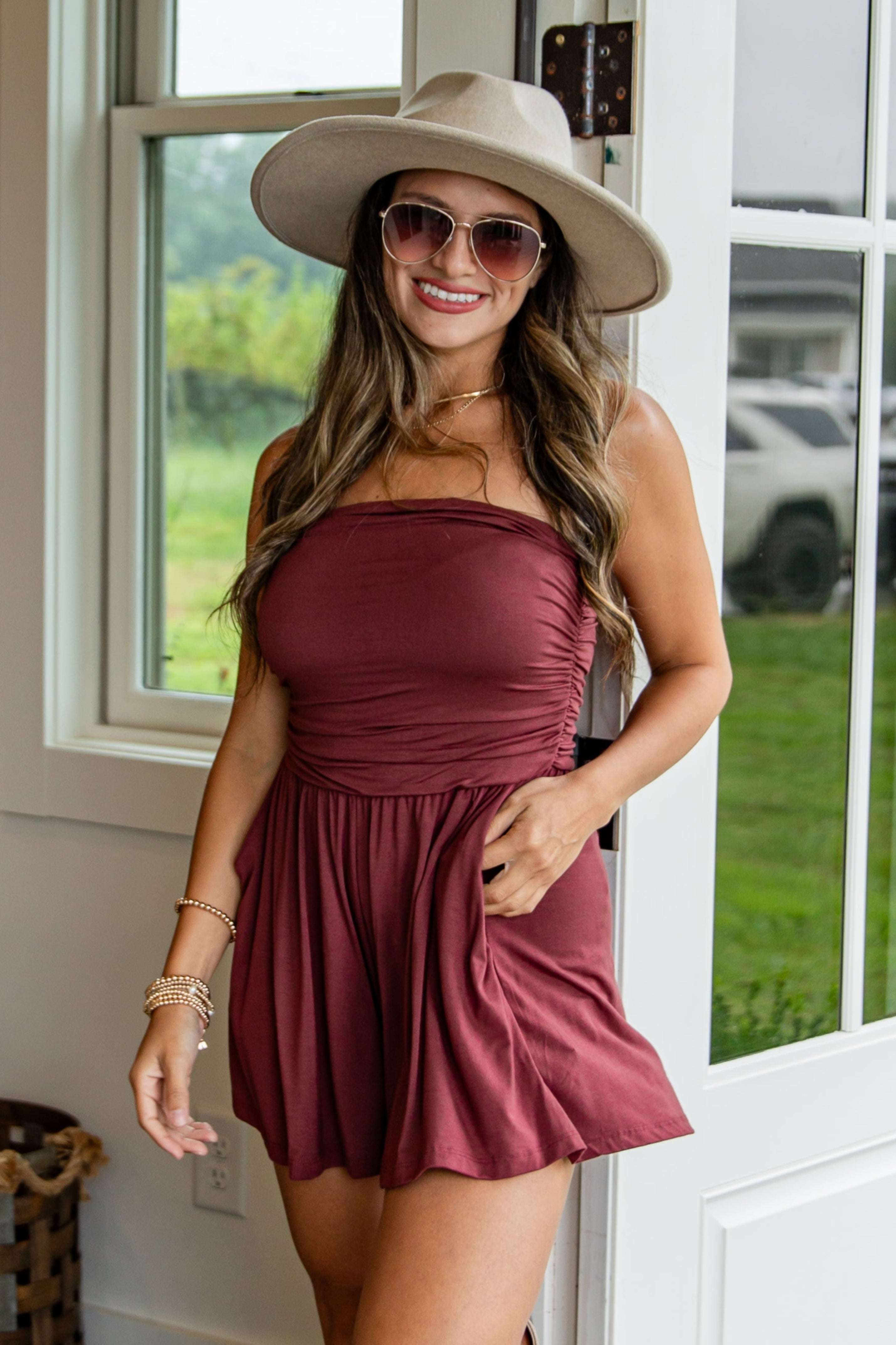 Woman in a burgundy dress and beige hat standing on a porch.
