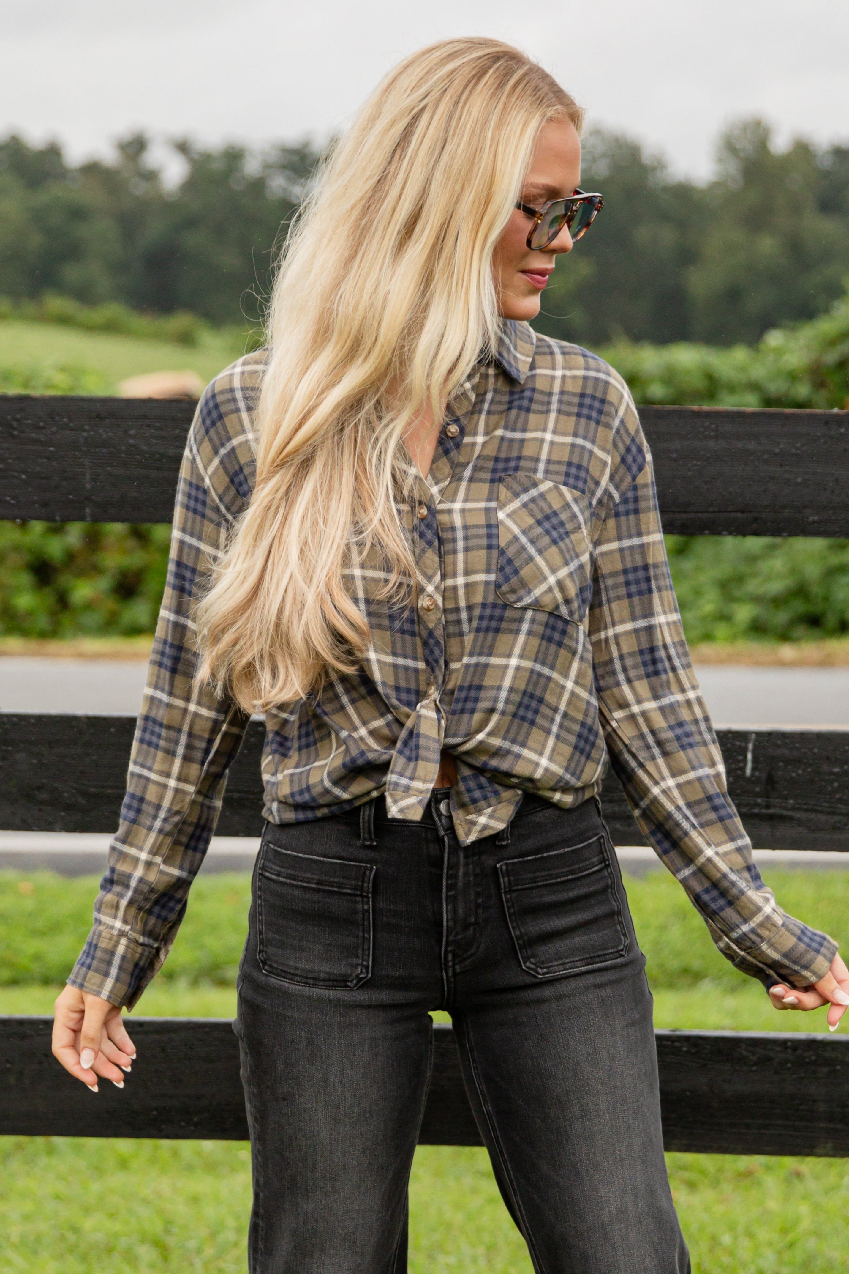 Woman wearing a plaid shirt and sunglasses standing in front of a wooden fence with greenery in the background