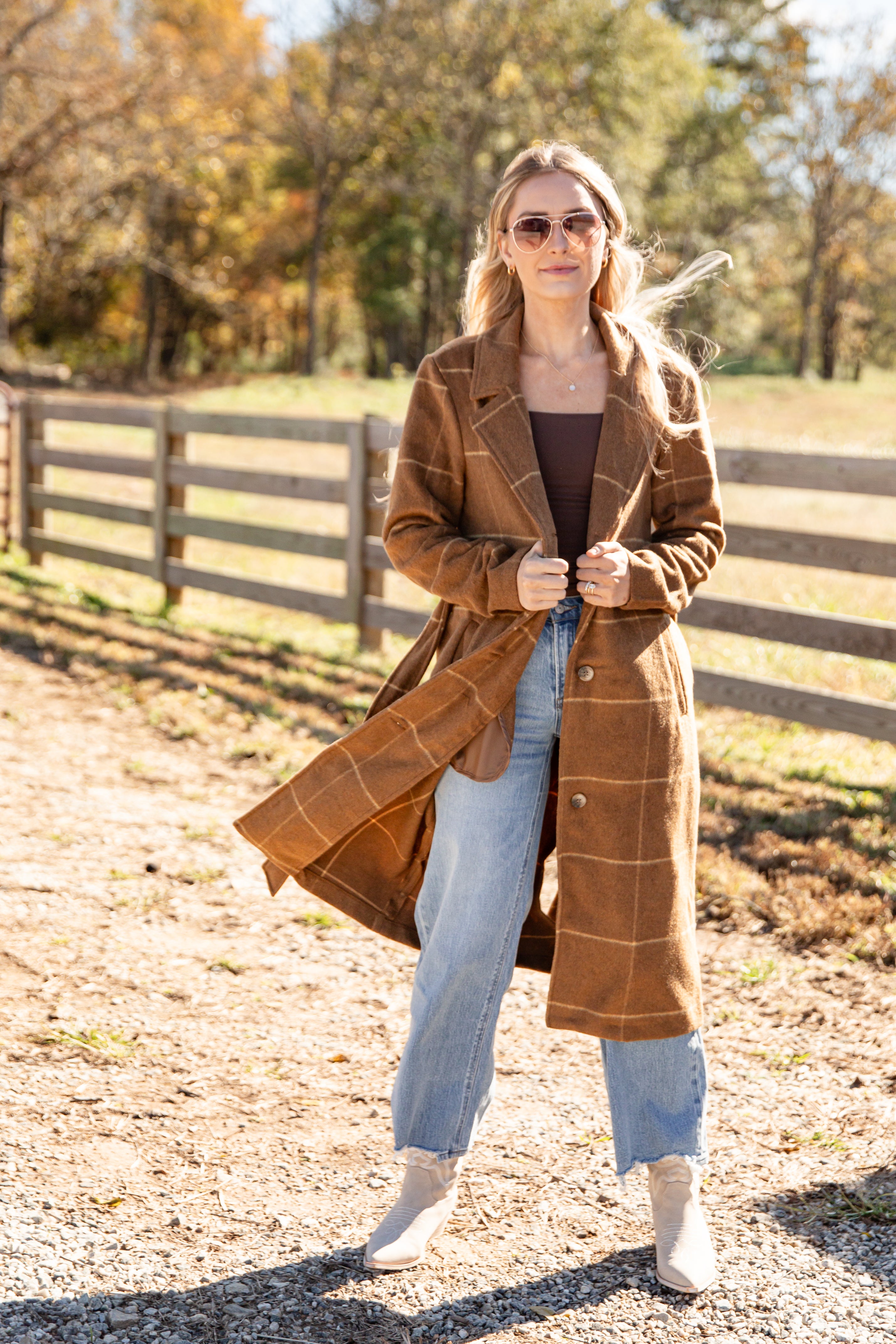 Woman wearing a brown plaid coat in an outdoor setting with trees and a fence.