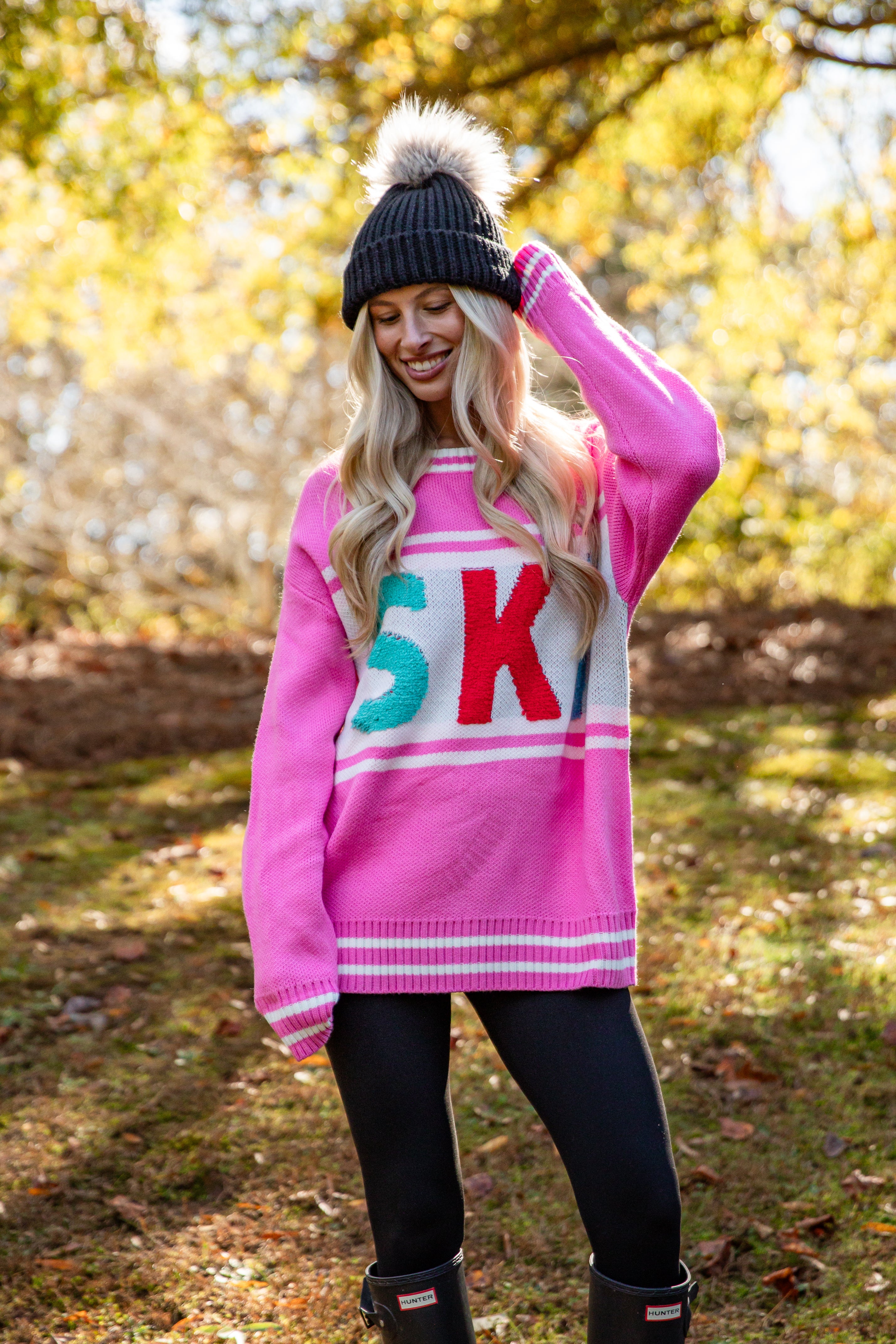 Woman wearing a pink sweater with colorful letters, standing outdoors in a natural setting.