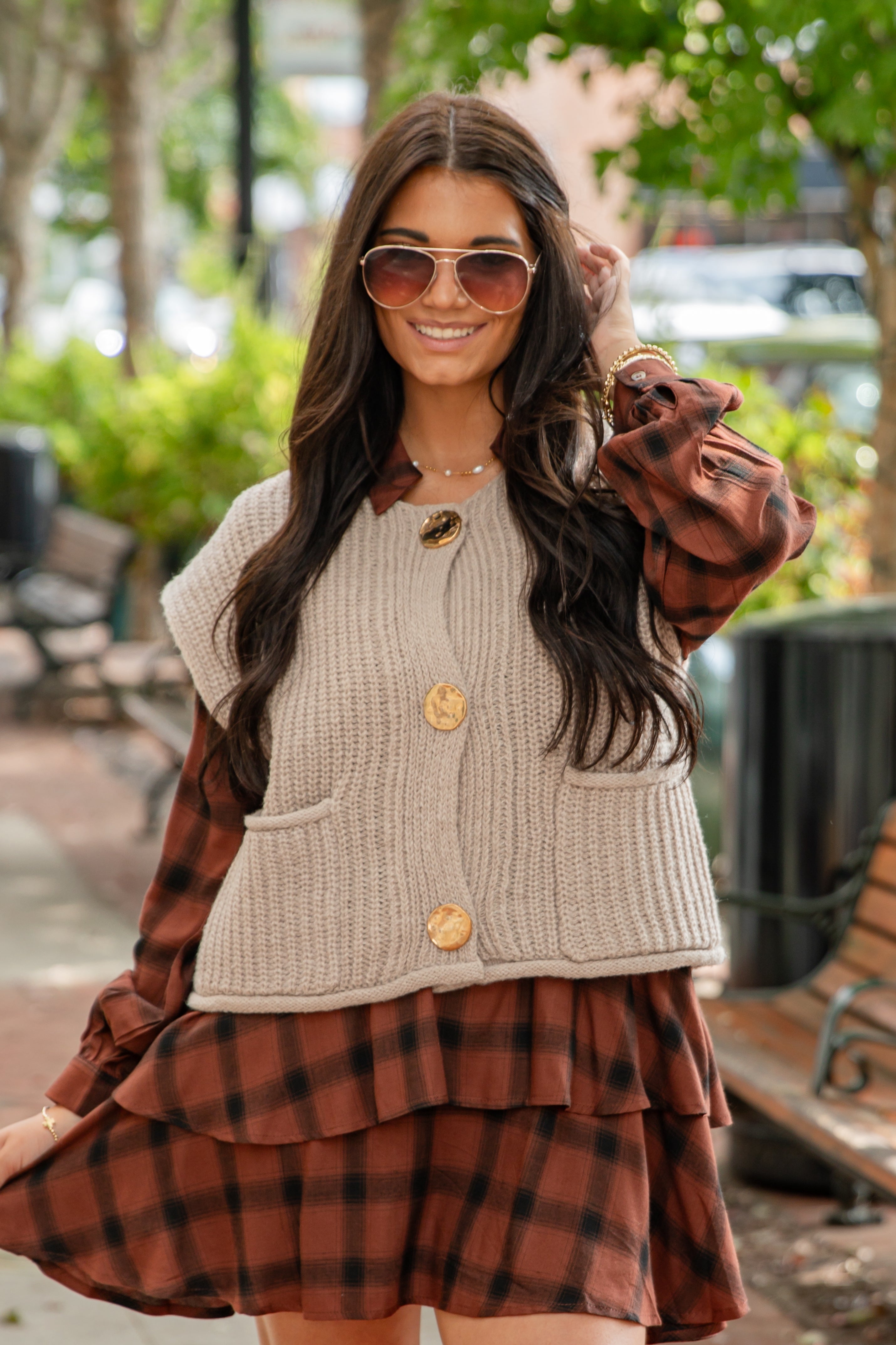 Woman wearing a plaid dress over a beige cardigan with sunglasses outdoors.