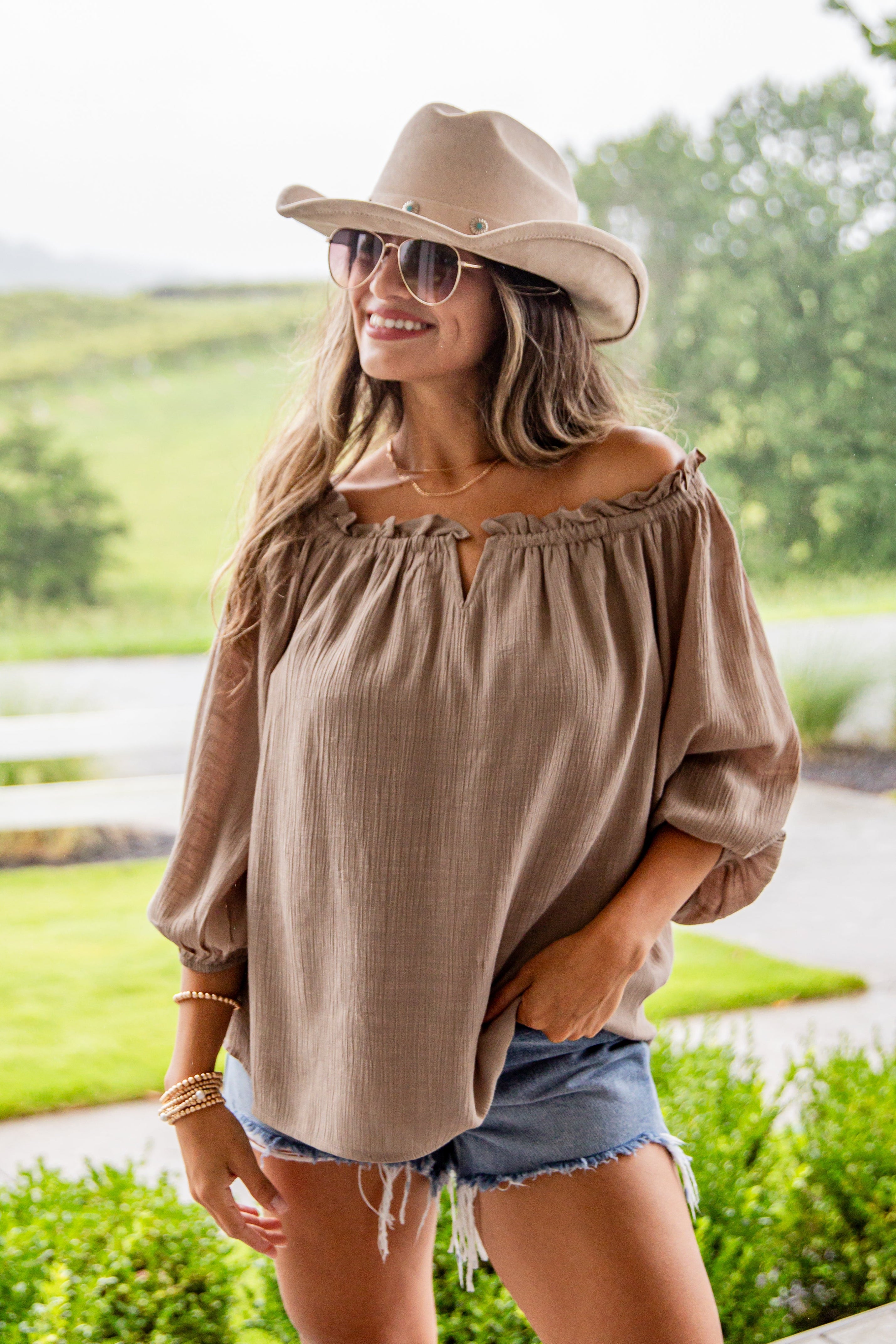 Woman wearing a beige off-shoulder top and denim shorts with sunglasses and a hat outdoors.