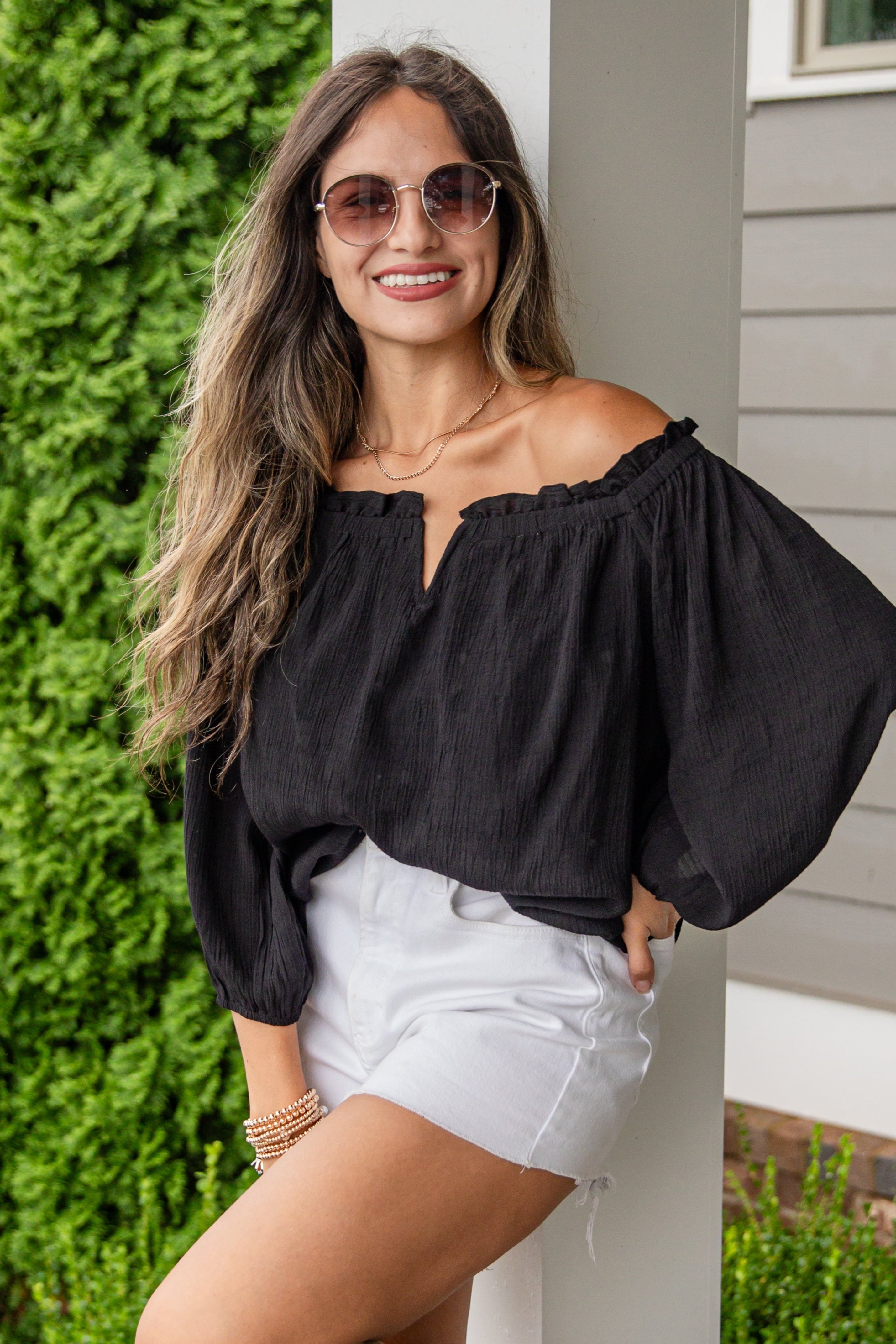 Woman in black top and white shorts standing on a porch with greenery in the background
