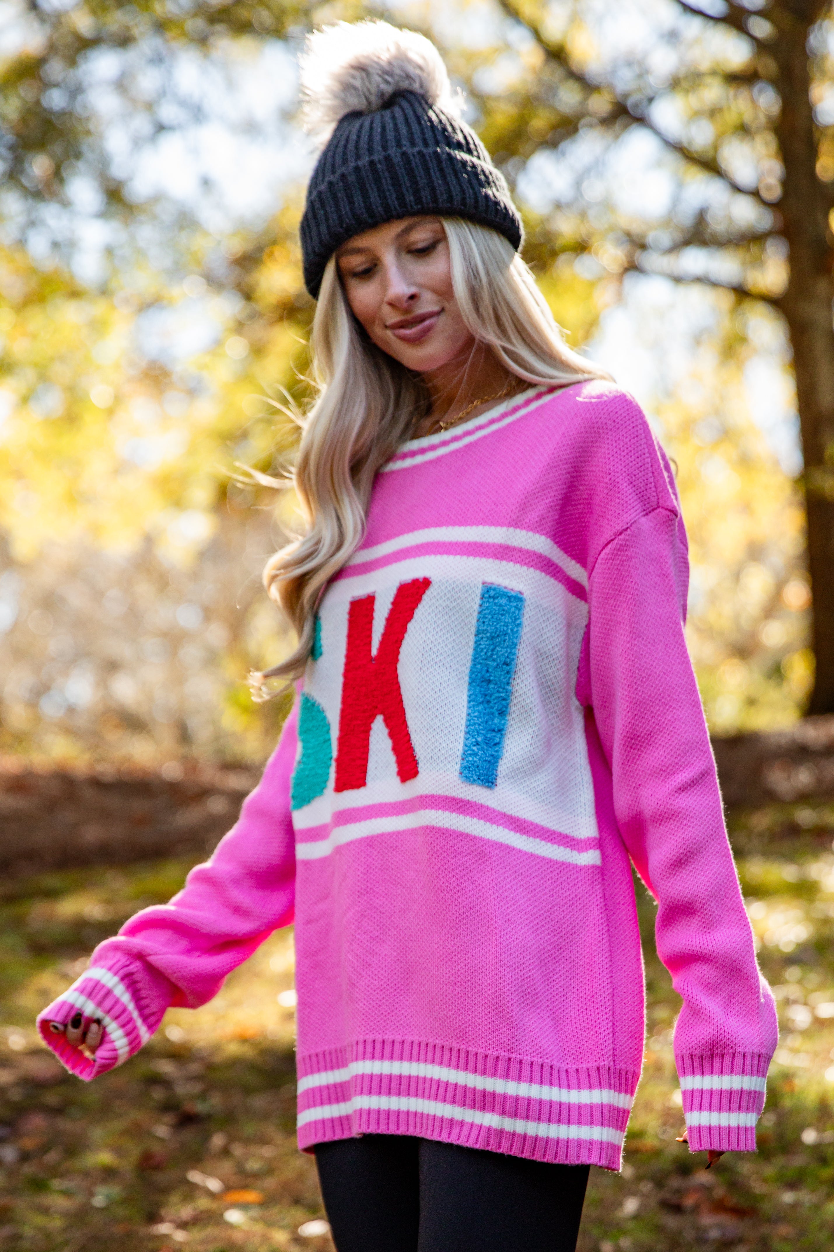 Woman wearing a pink sweater with colorful letters and a black beanie outdoors.
