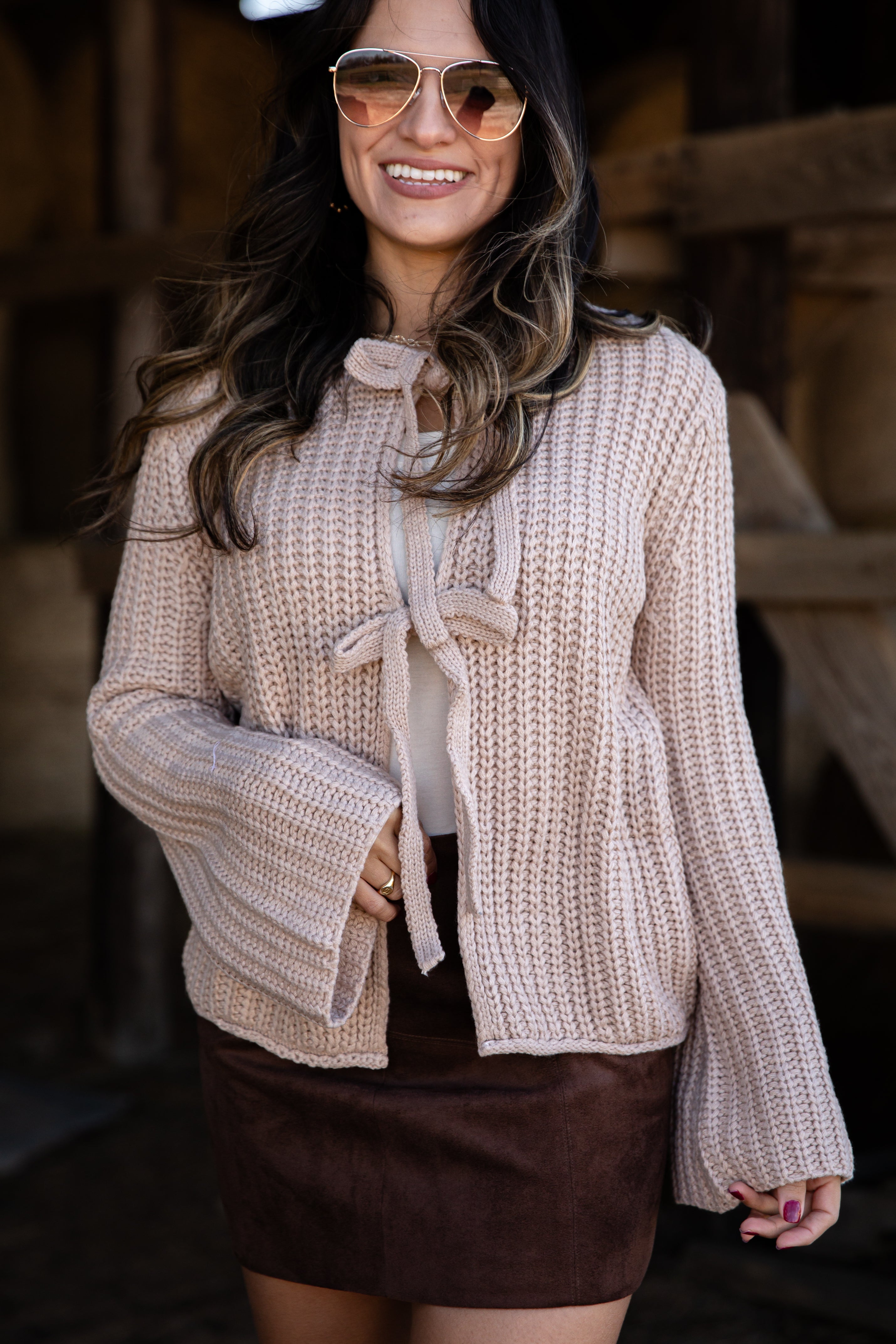 Woman wearing a beige knitted jacket with a bow tie, smiling outdoors.