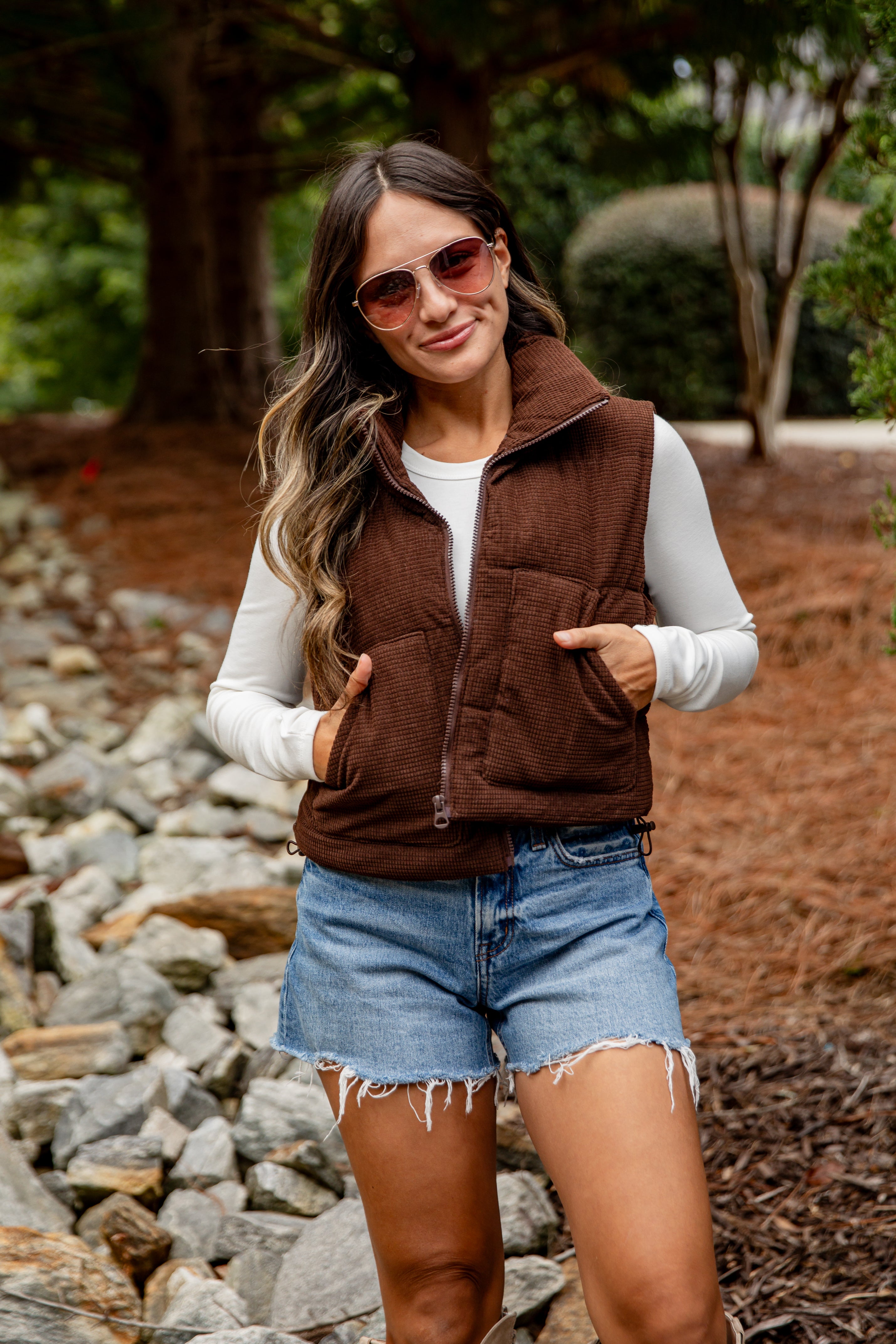 Woman wearing a brown vest, white shirt, and denim shorts outdoors.