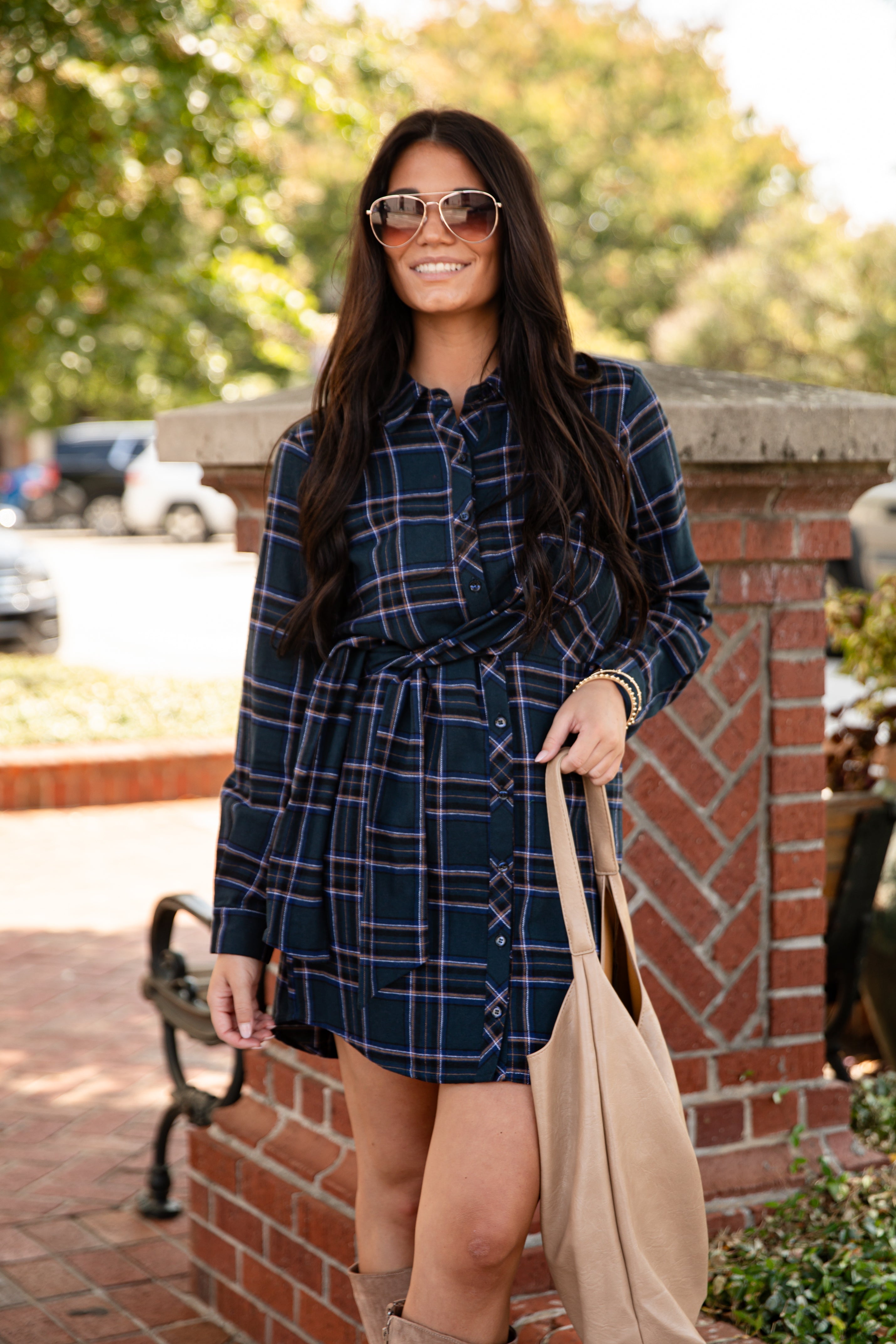 Woman wearing a plaid dress with sunglasses and a beige bag outdoors.