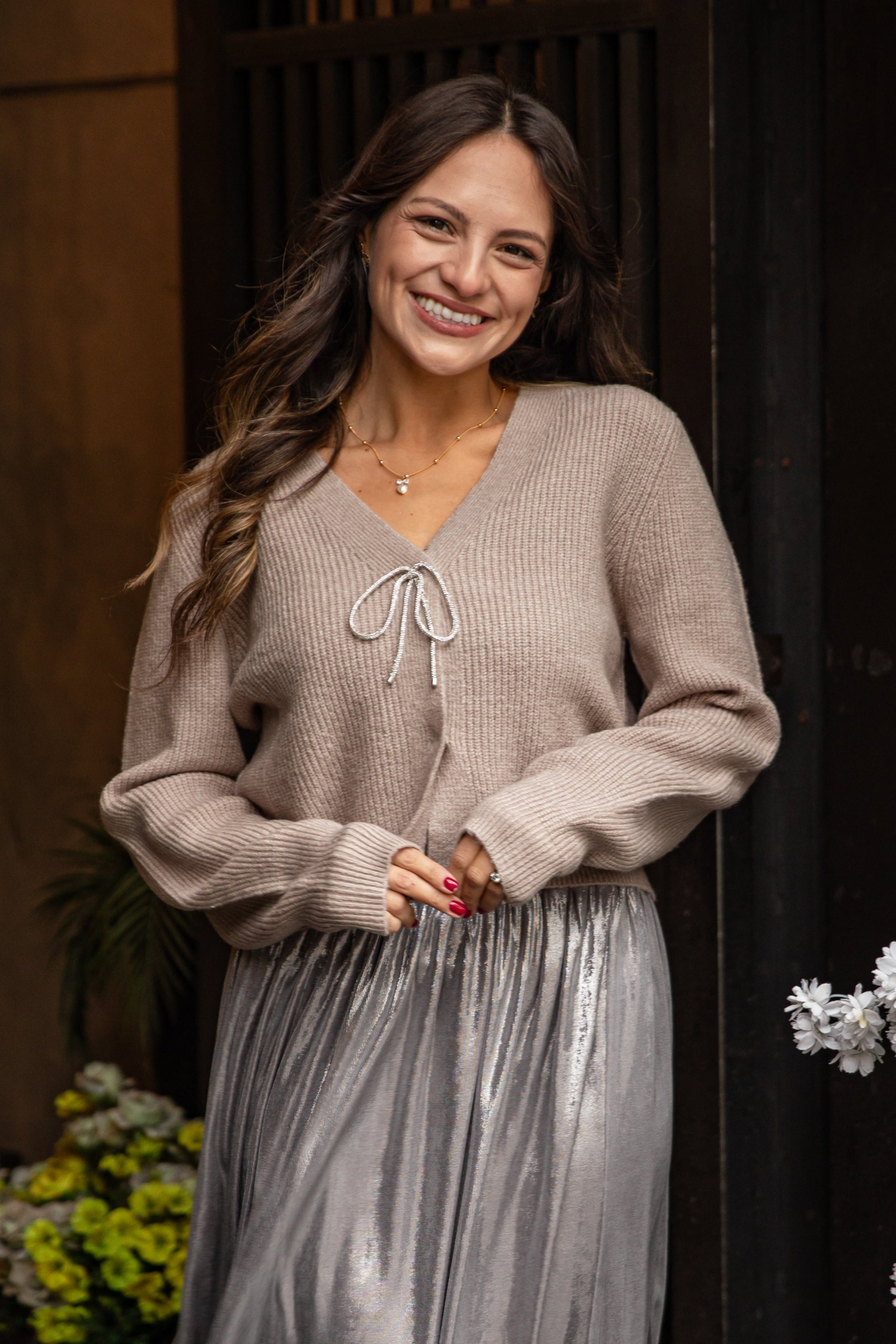 Woman wearing a beige sweater and silver skirt standing in front of a decorative door with flowers.