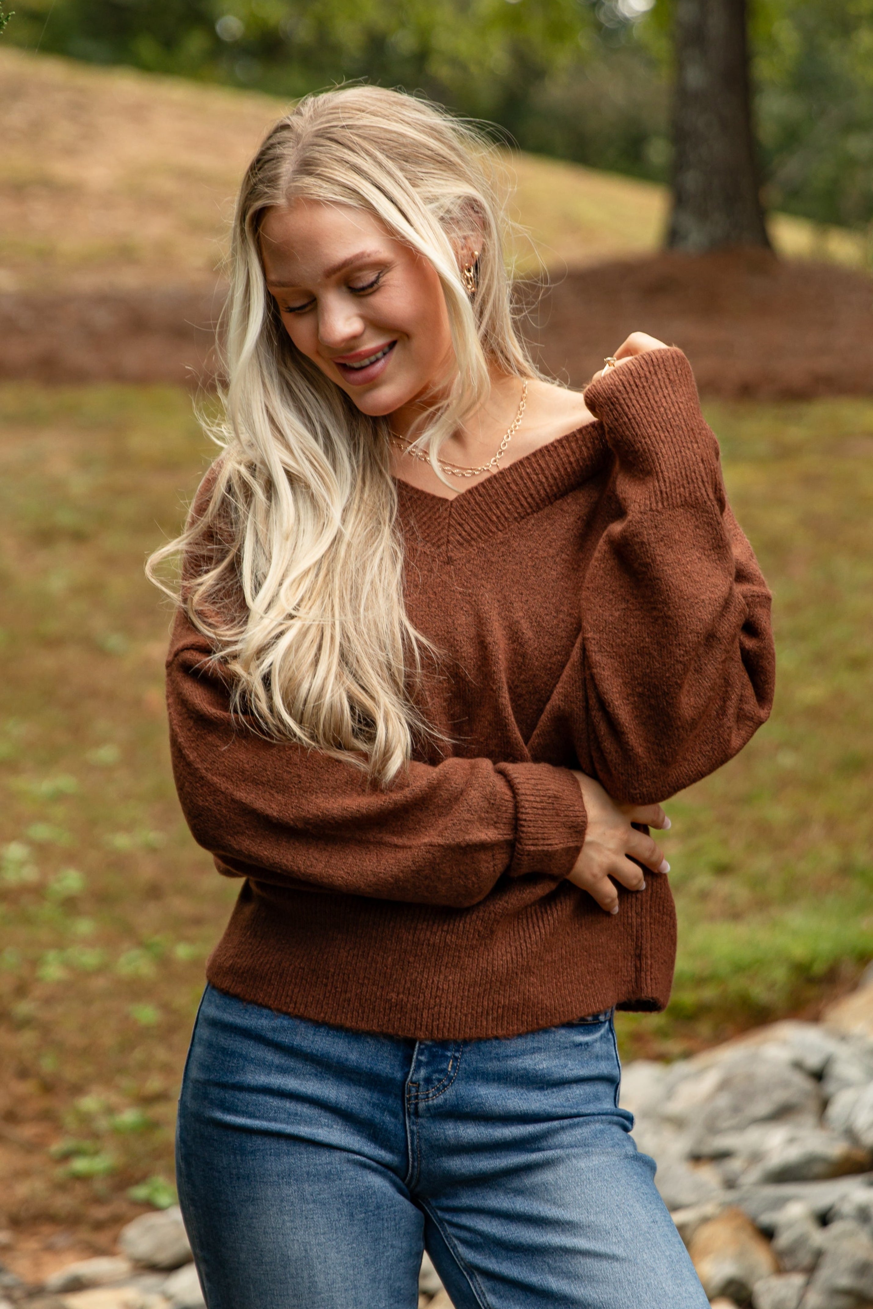Woman wearing a brown sweater and blue jeans standing outdoors with trees and rocks in the background