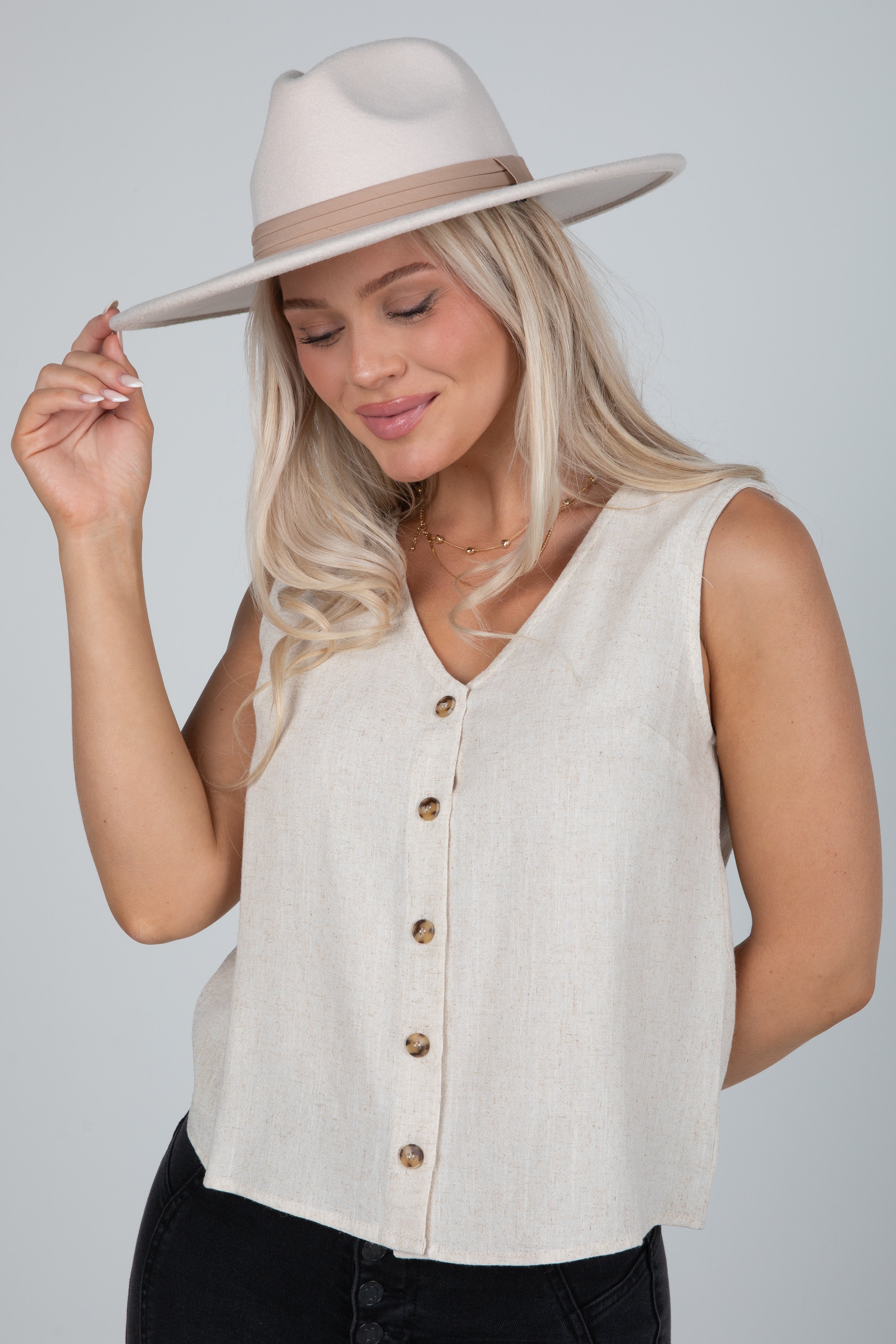 Woman wearing a beige sleeveless button-up top and hat against a plain background