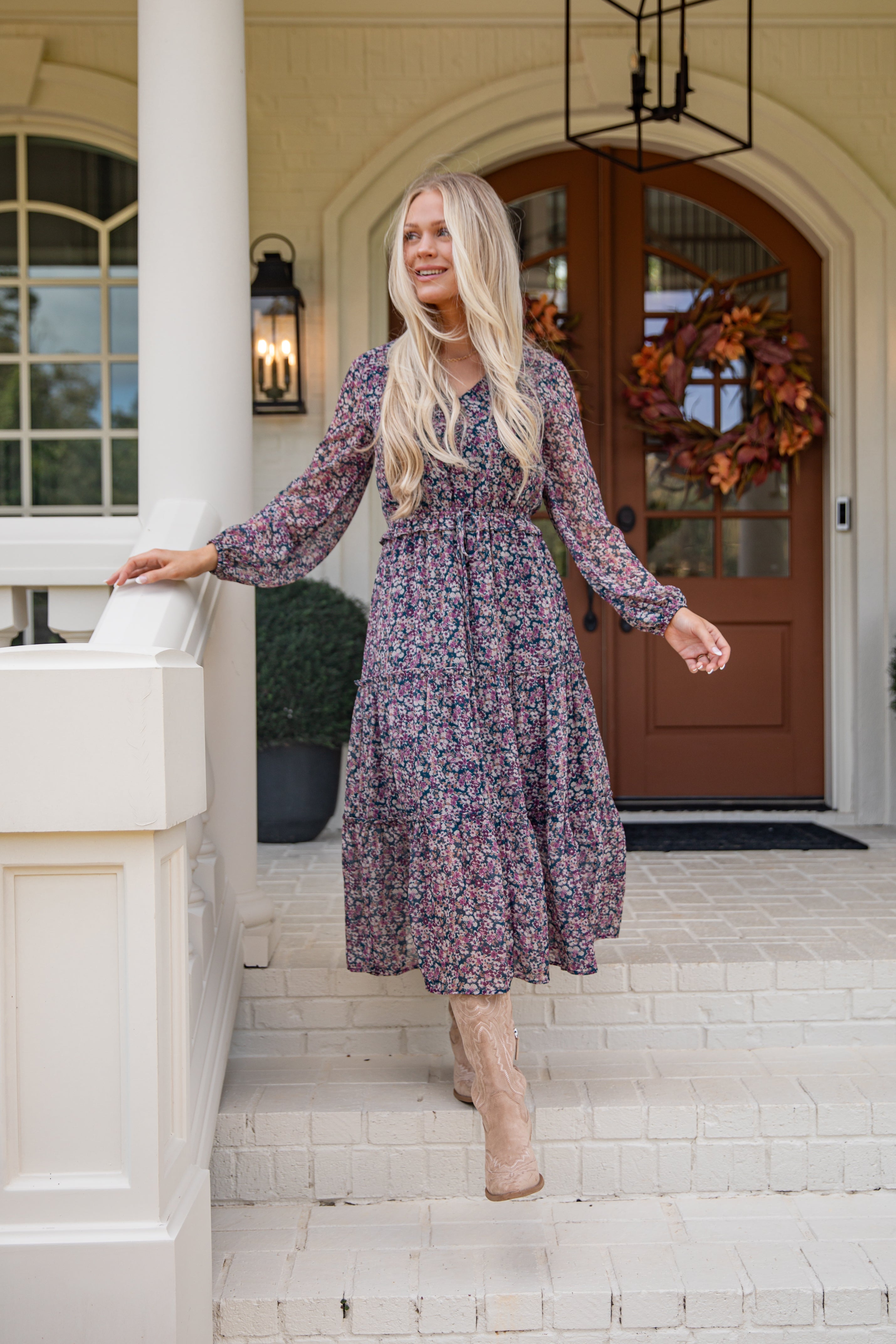 Woman in a floral dress standing on a porch with a wreath in the background