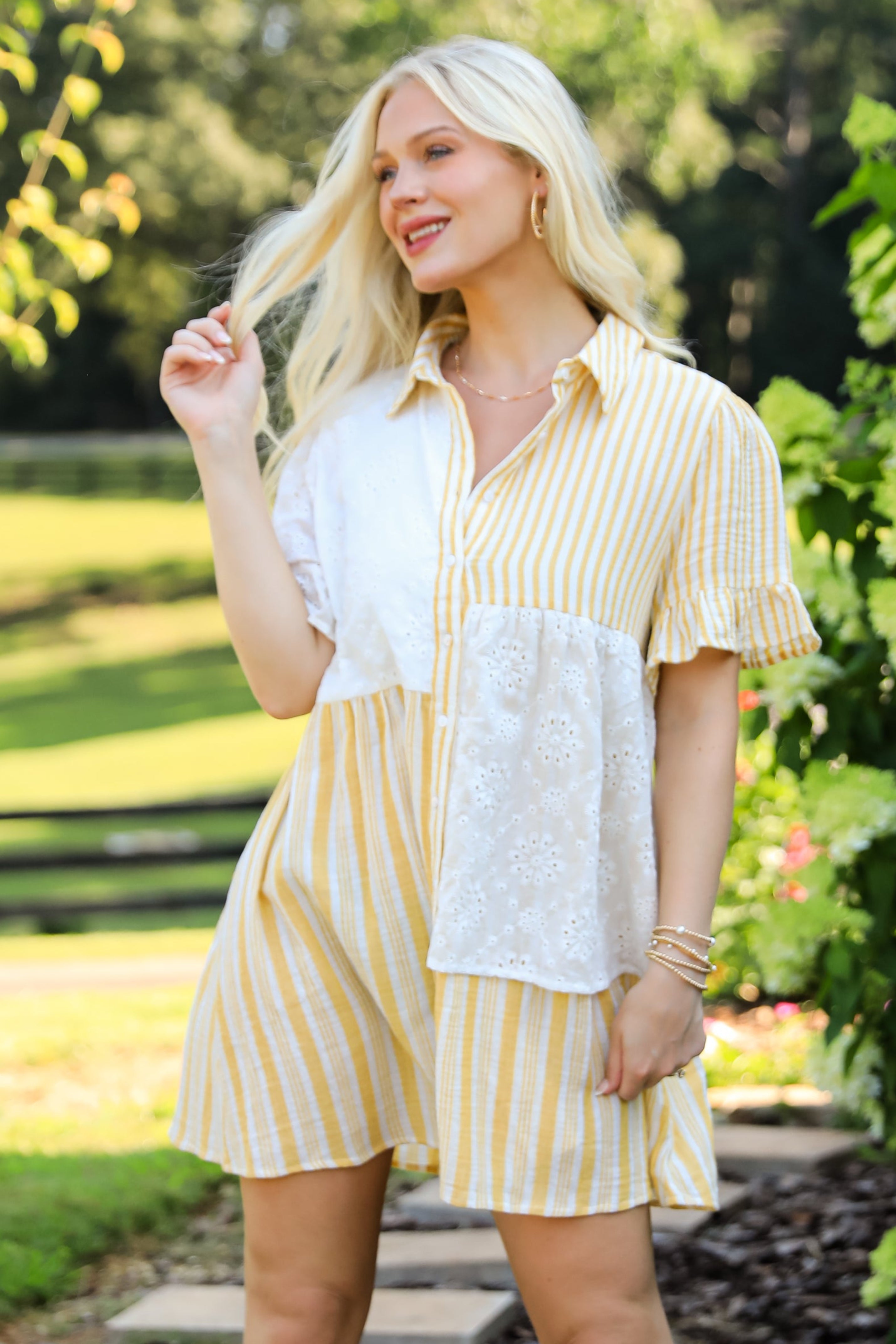 Woman in a yellow and white outfit standing on a wooden path with greenery around.