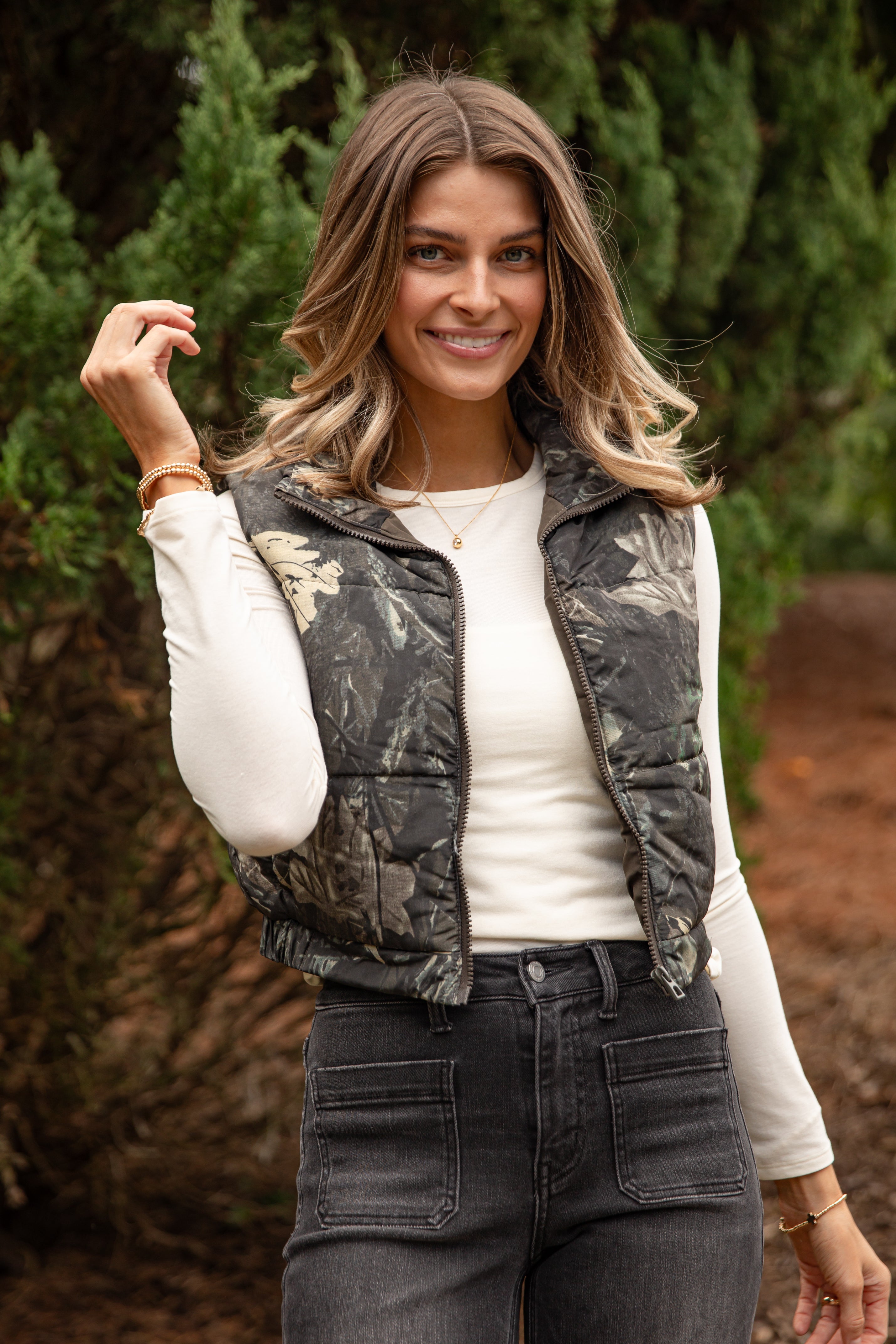 Woman wearing a camouflage vest over a white shirt with trees in the background