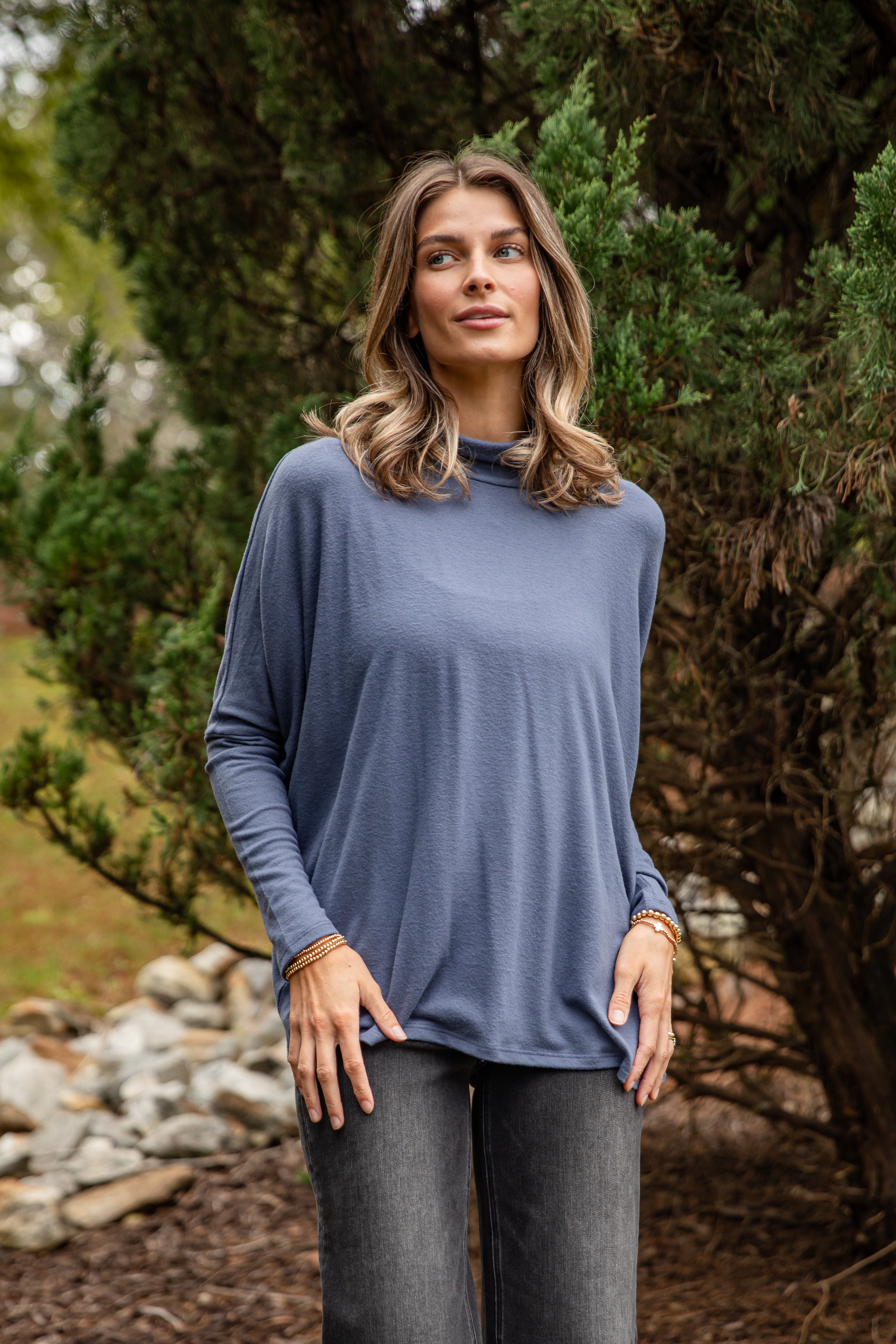 Woman wearing a blue long-sleeve top and gray pants standing outdoors with greenery in the background
