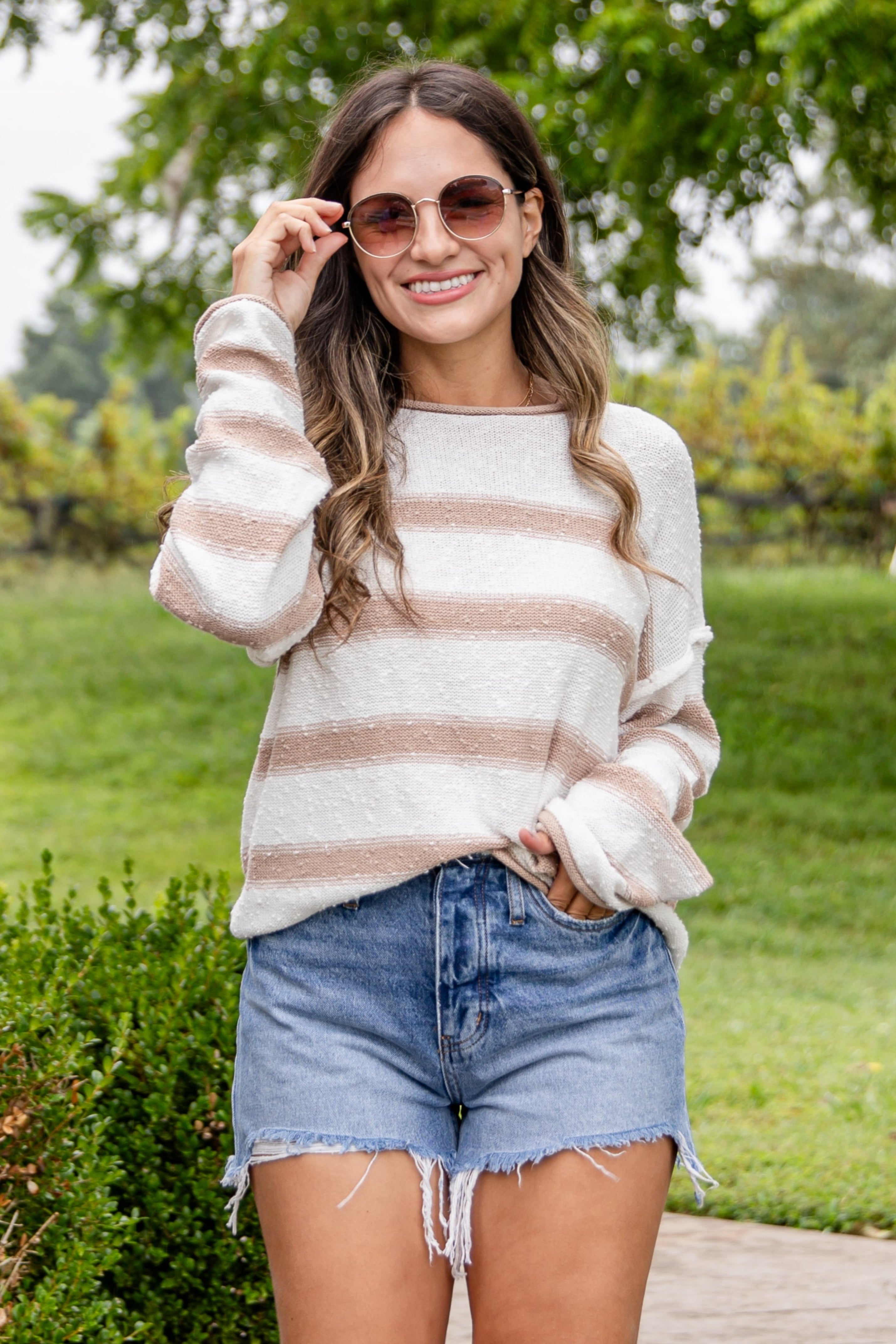Woman in a striped sweater and denim shorts walking outdoors with greenery in the background