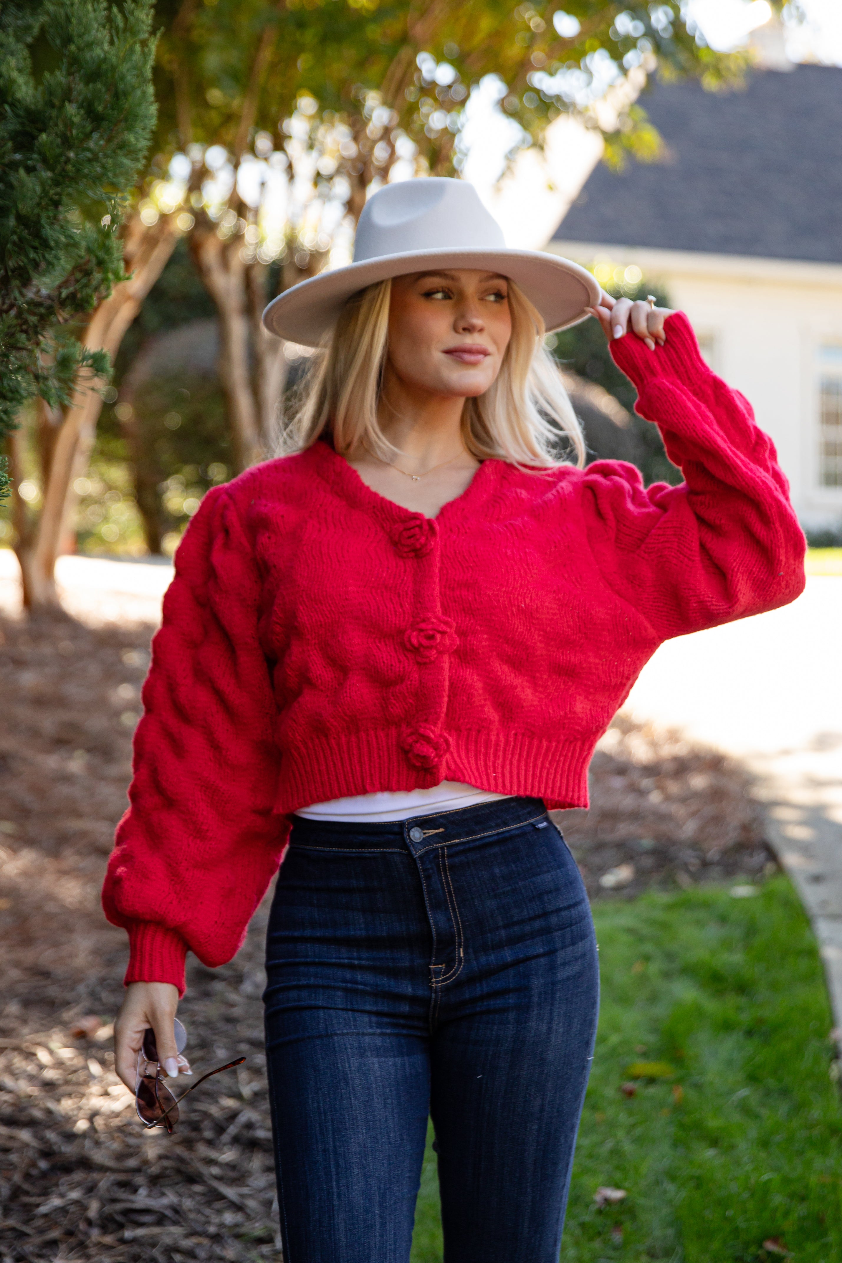 Woman wearing a red cardigan, white shirt, blue jeans, and a wide-brimmed hat outdoors.