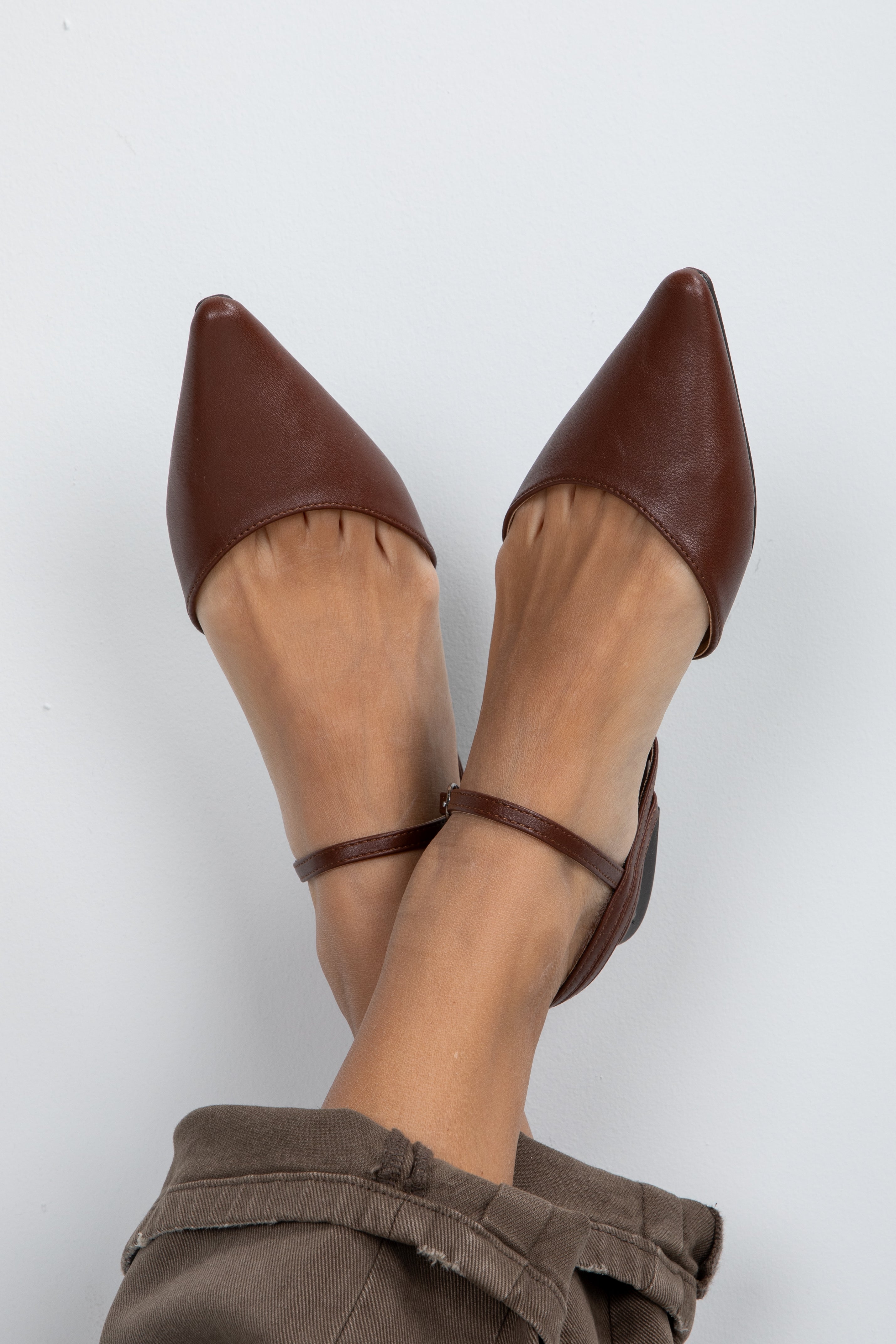Brown pointed-toe shoes with ankle straps worn by a person on a white background