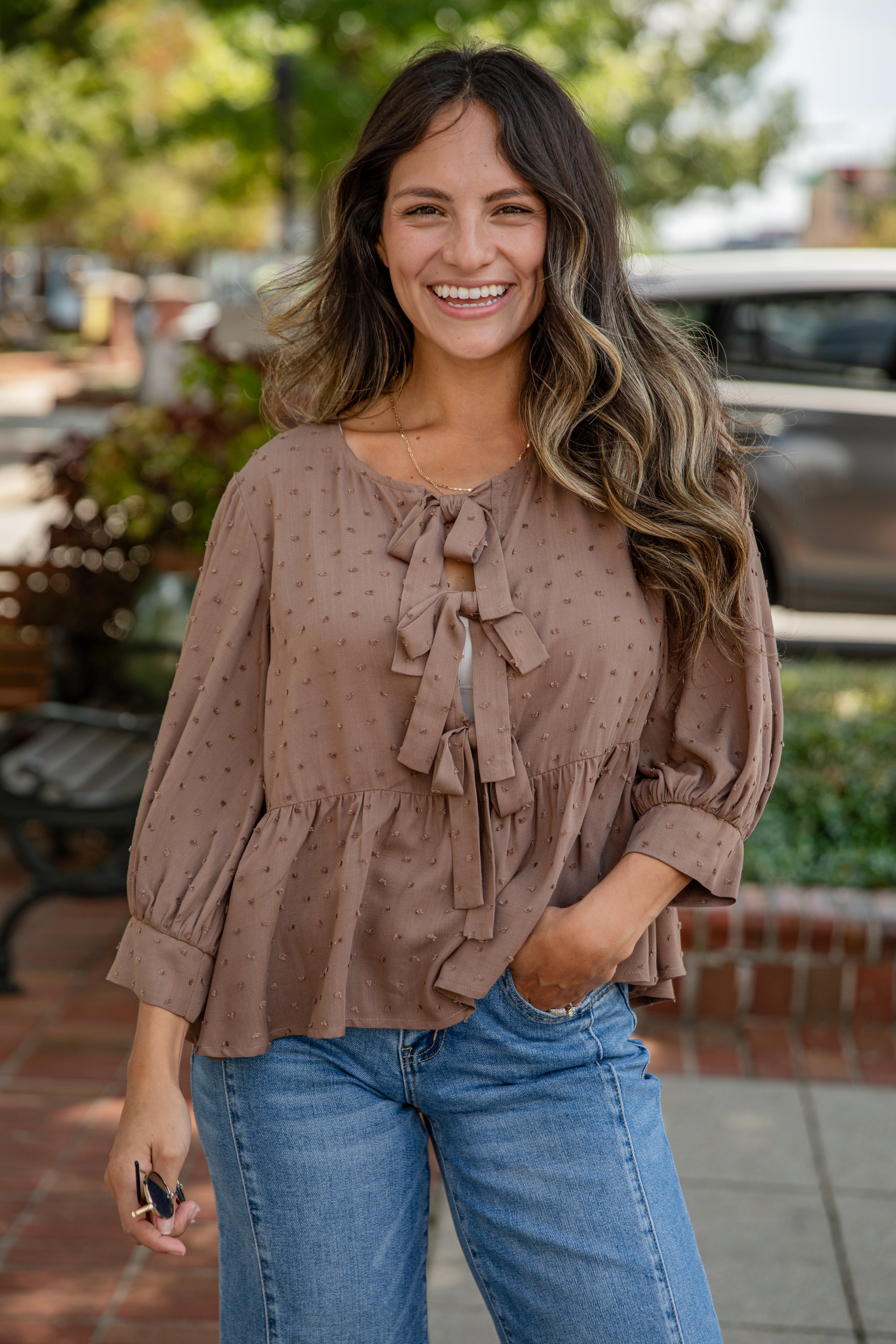 Woman wearing a brown blouse with ruffled sleeves and blue jeans, standing outdoors.